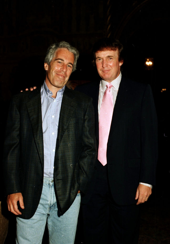 Two men pose together, one in a casual blazer and shirt, the other in a suit with a pink tie. They stand indoors, facing the camera