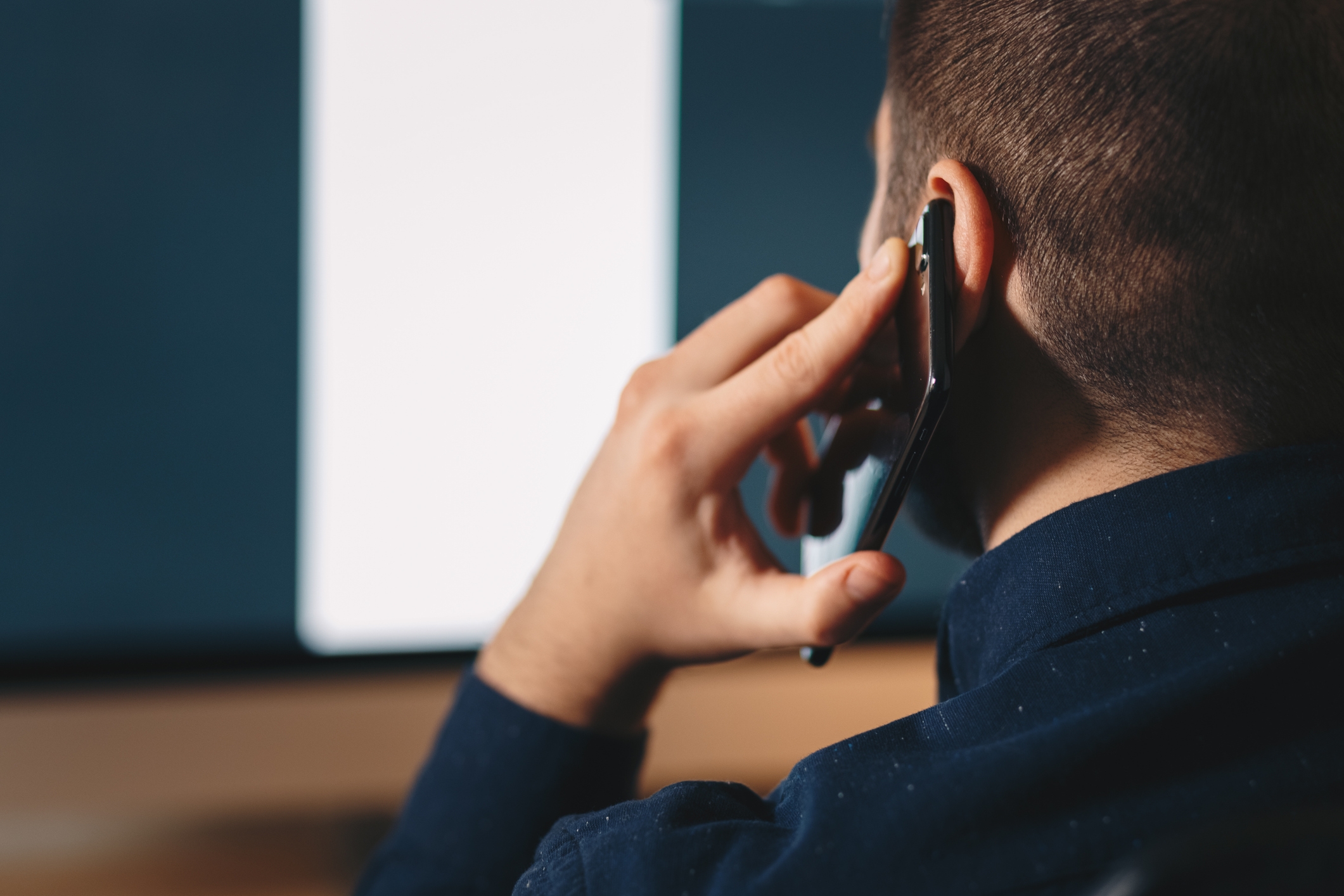Person holding a smartphone to their ear while looking at a computer screen, suggesting they are on a phone call