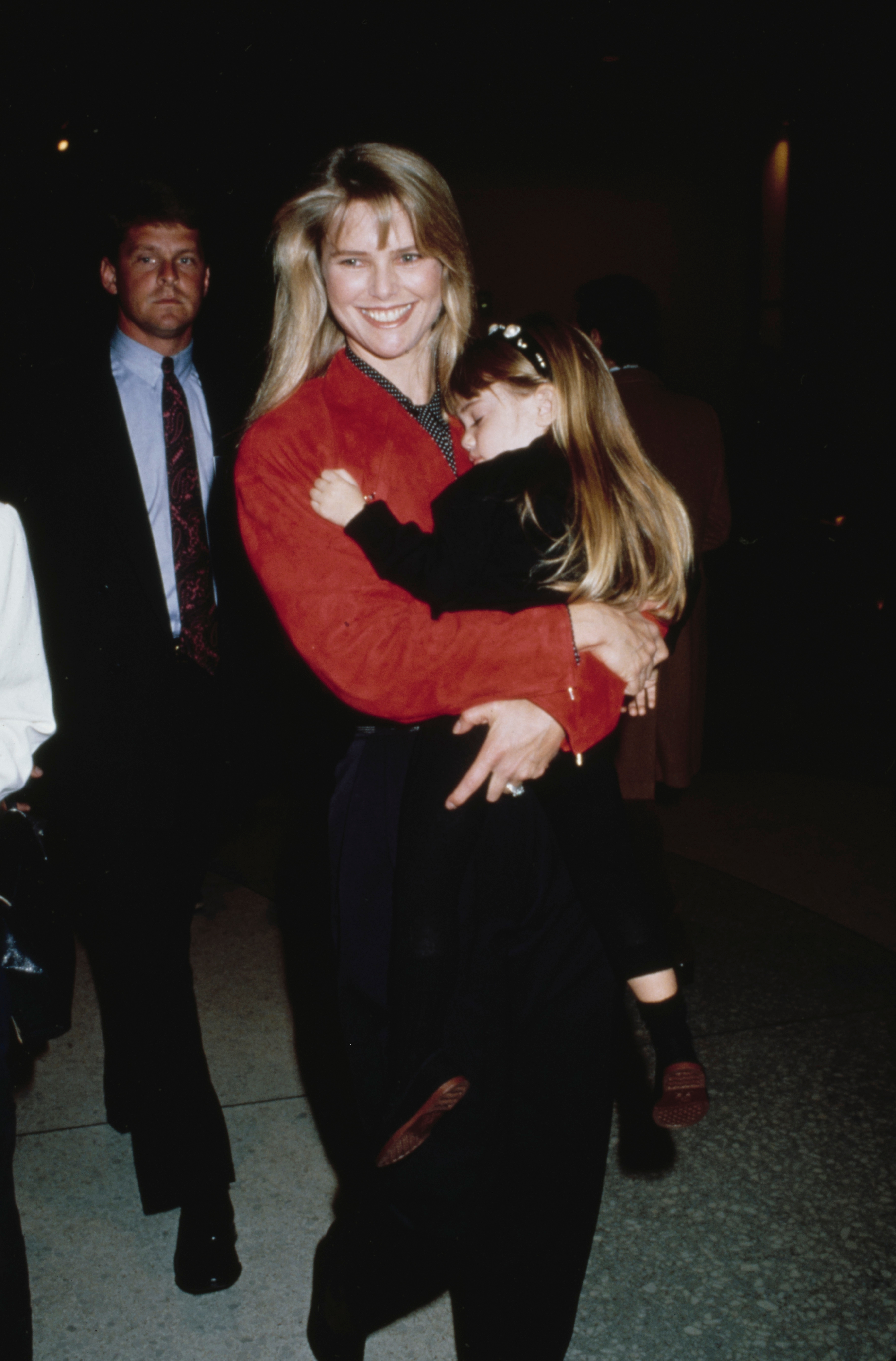 Woman smiling, holding a child in her arms. People are in the background, and the setting appears candid and casual