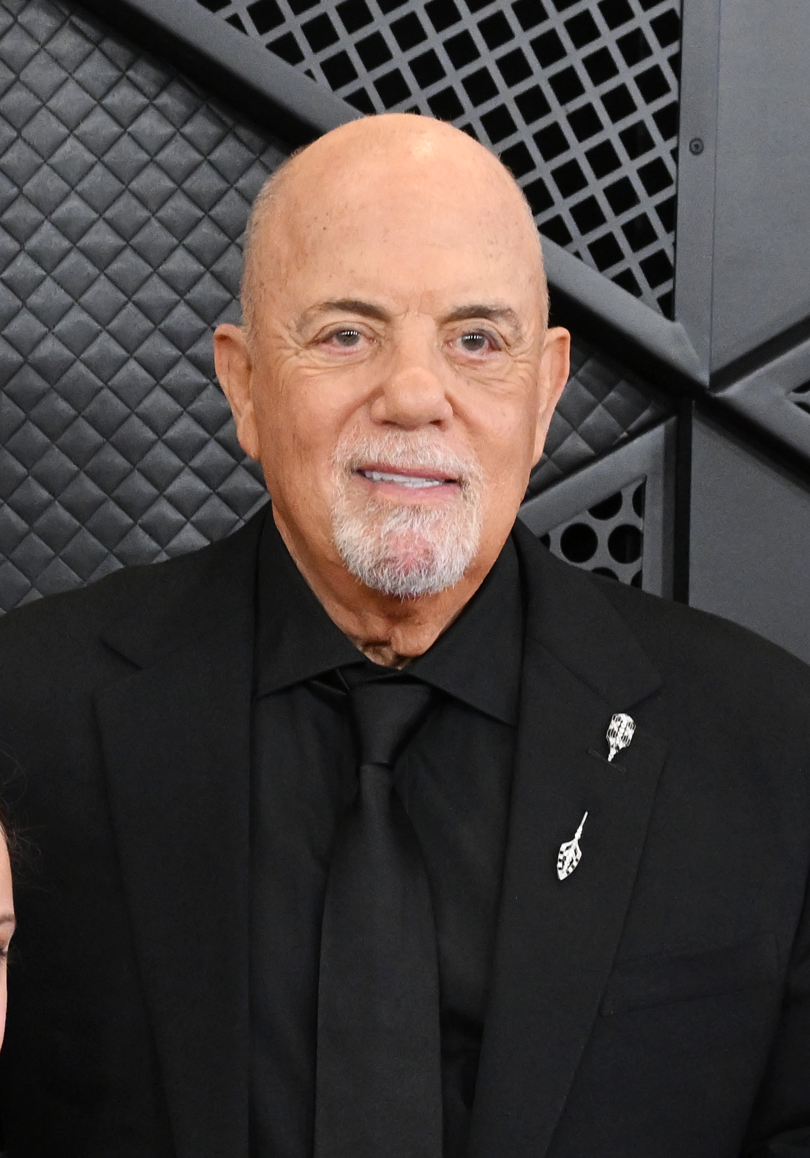 A person in a formal black suit and tie with pin accessories stands against an embossed backdrop, smiling slightly