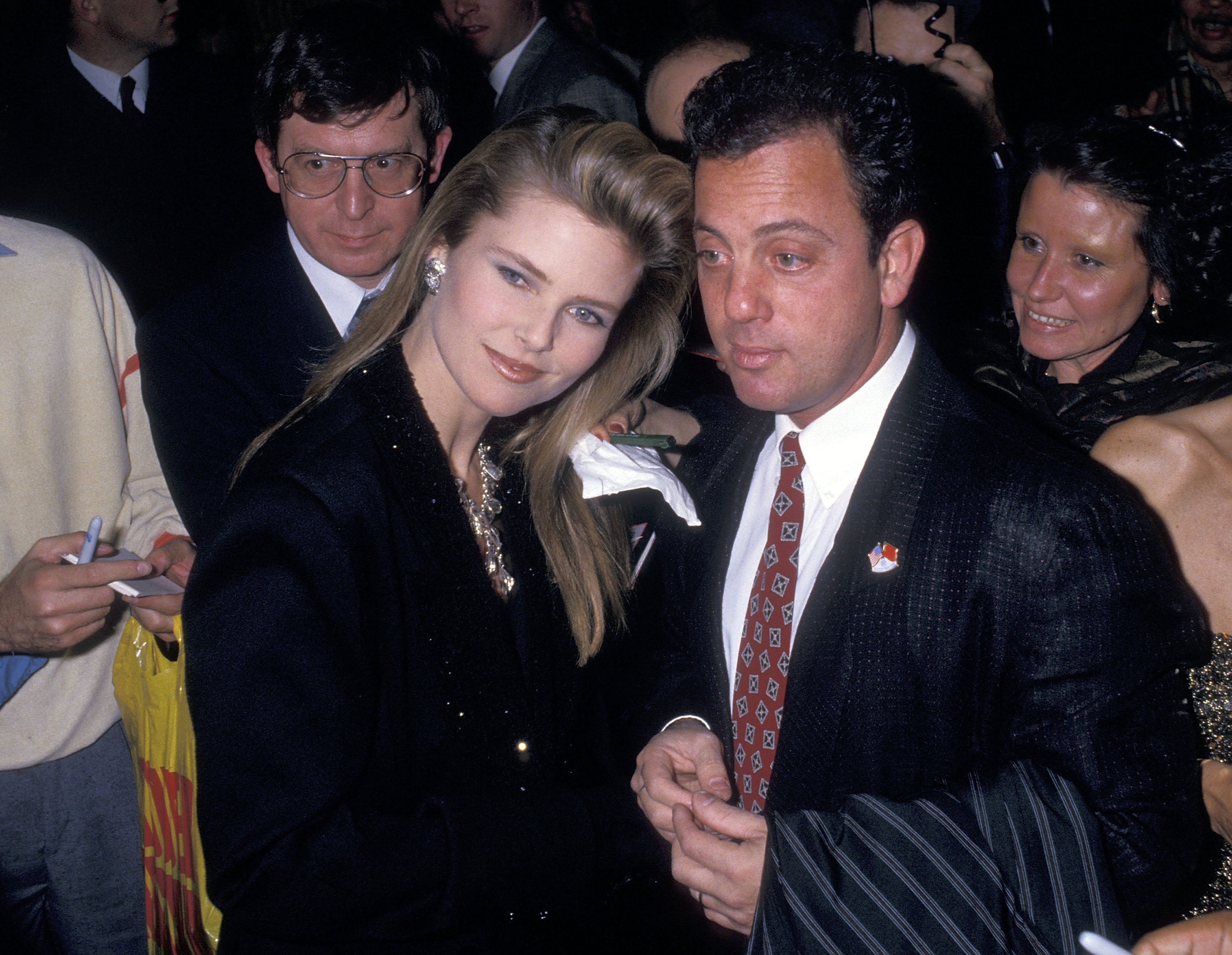 Two people at a formal event, one in a dark suit and tie, the other in an elegant black outfit. They are surrounded by onlookers