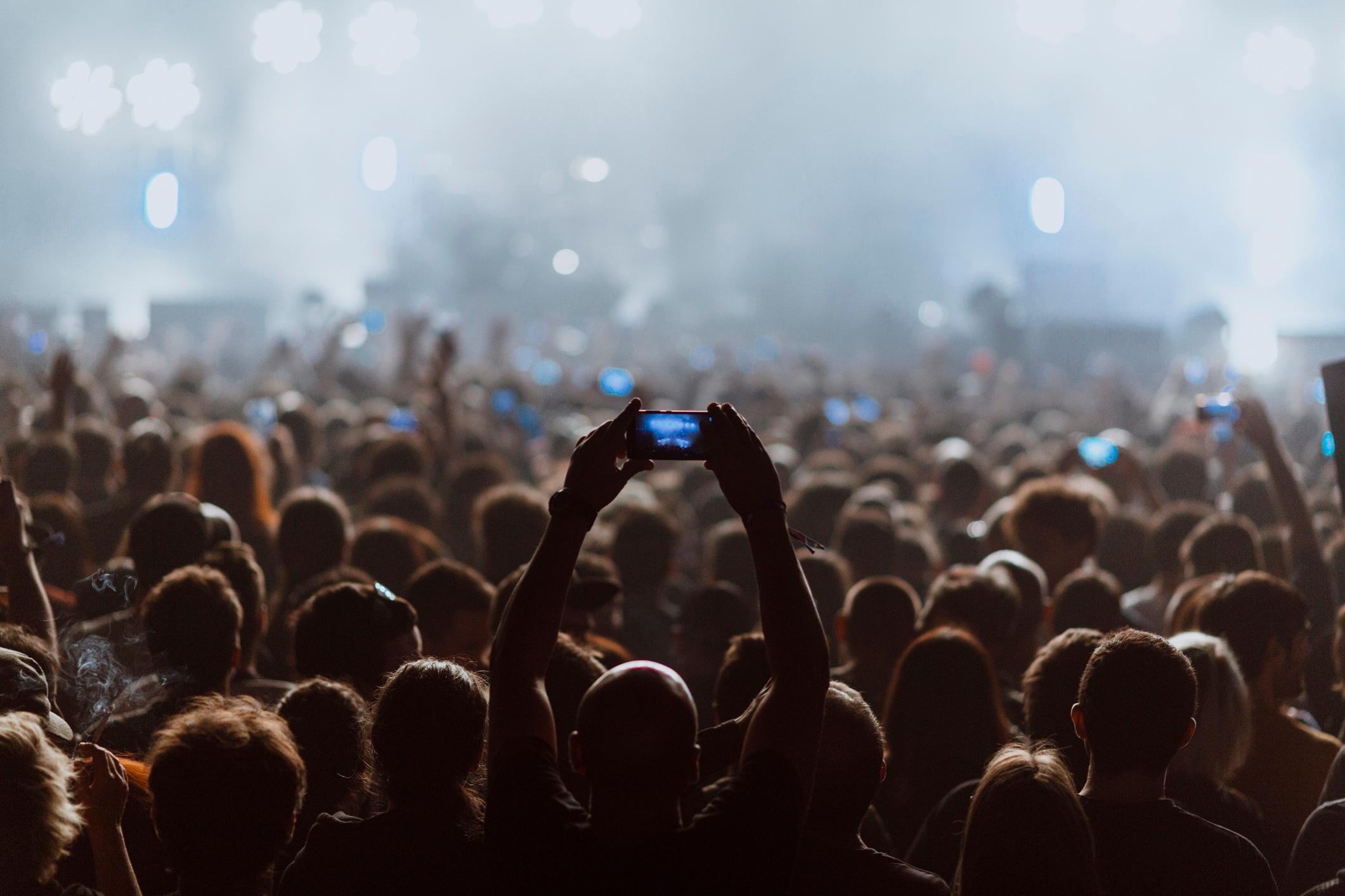 Audience at a concert, many holding up phones to capture the performance on stage. Bright lights illuminate the crowd in the background