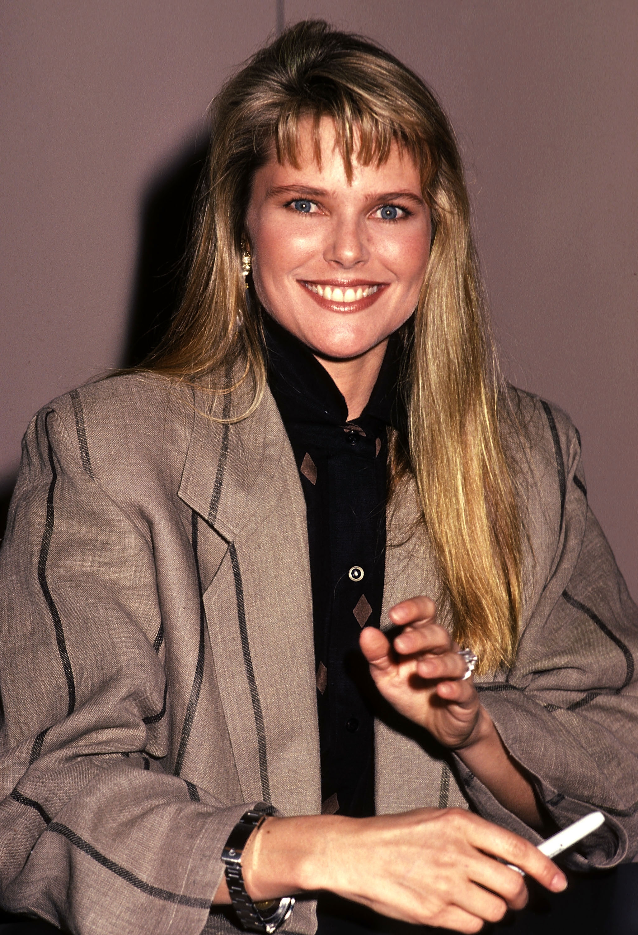 Person smiling, wearing a tailored blazer over a blouse. They are indoors and holding a pen, possibly at an event or interview