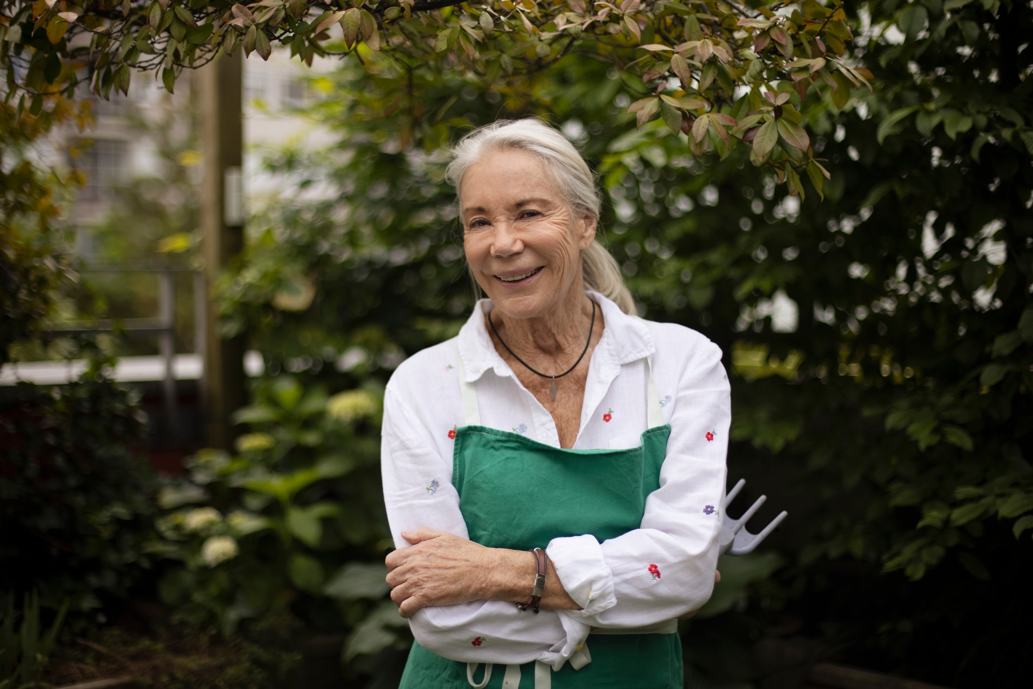 En eldre kvinne står i en hage, smiler, iført en hvit skjorte med et grønt forkle