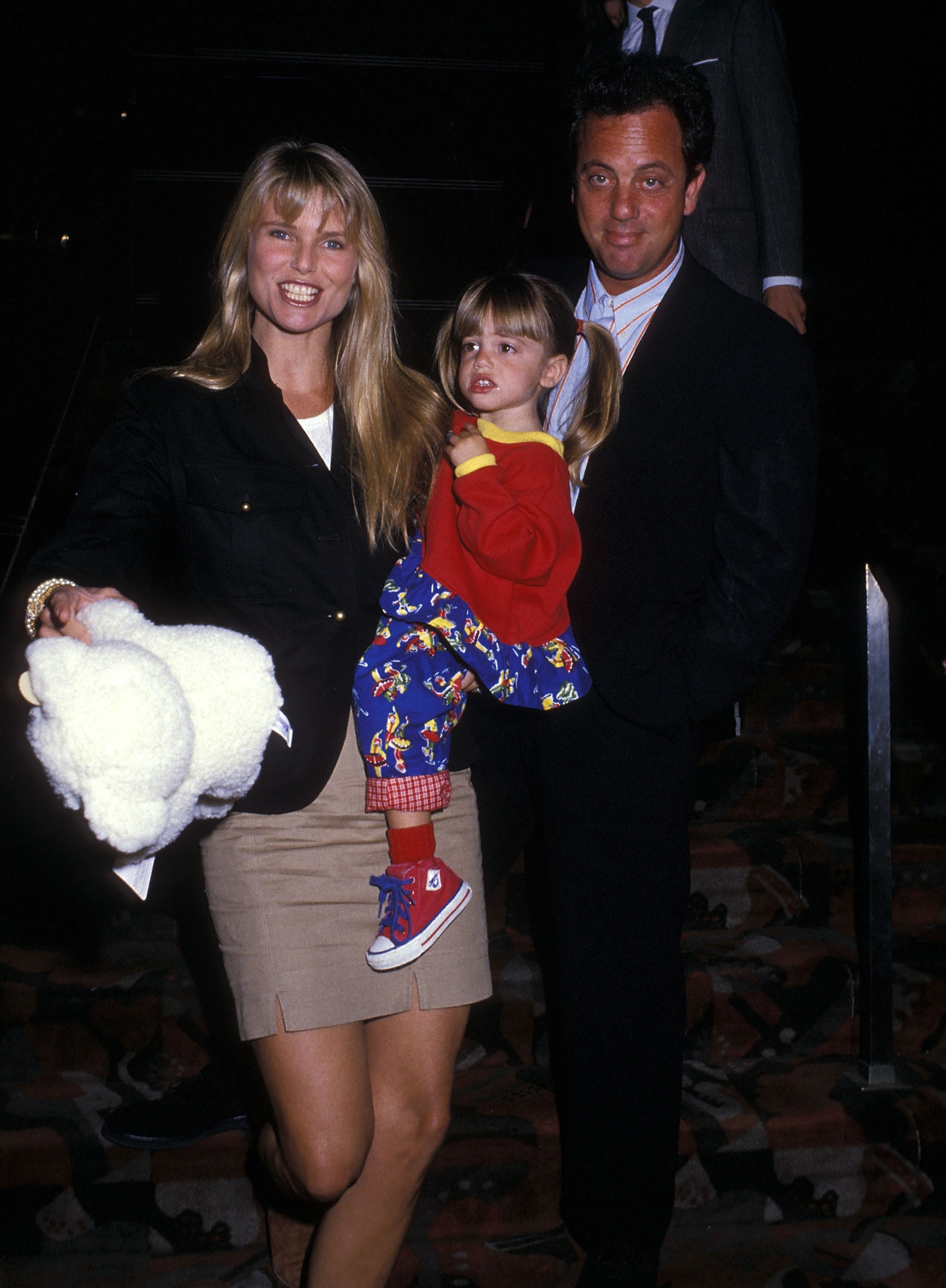 A family poses together; a woman in a blazer and skirt, a man in a suit, and a child in colorful overalls and red sneakers