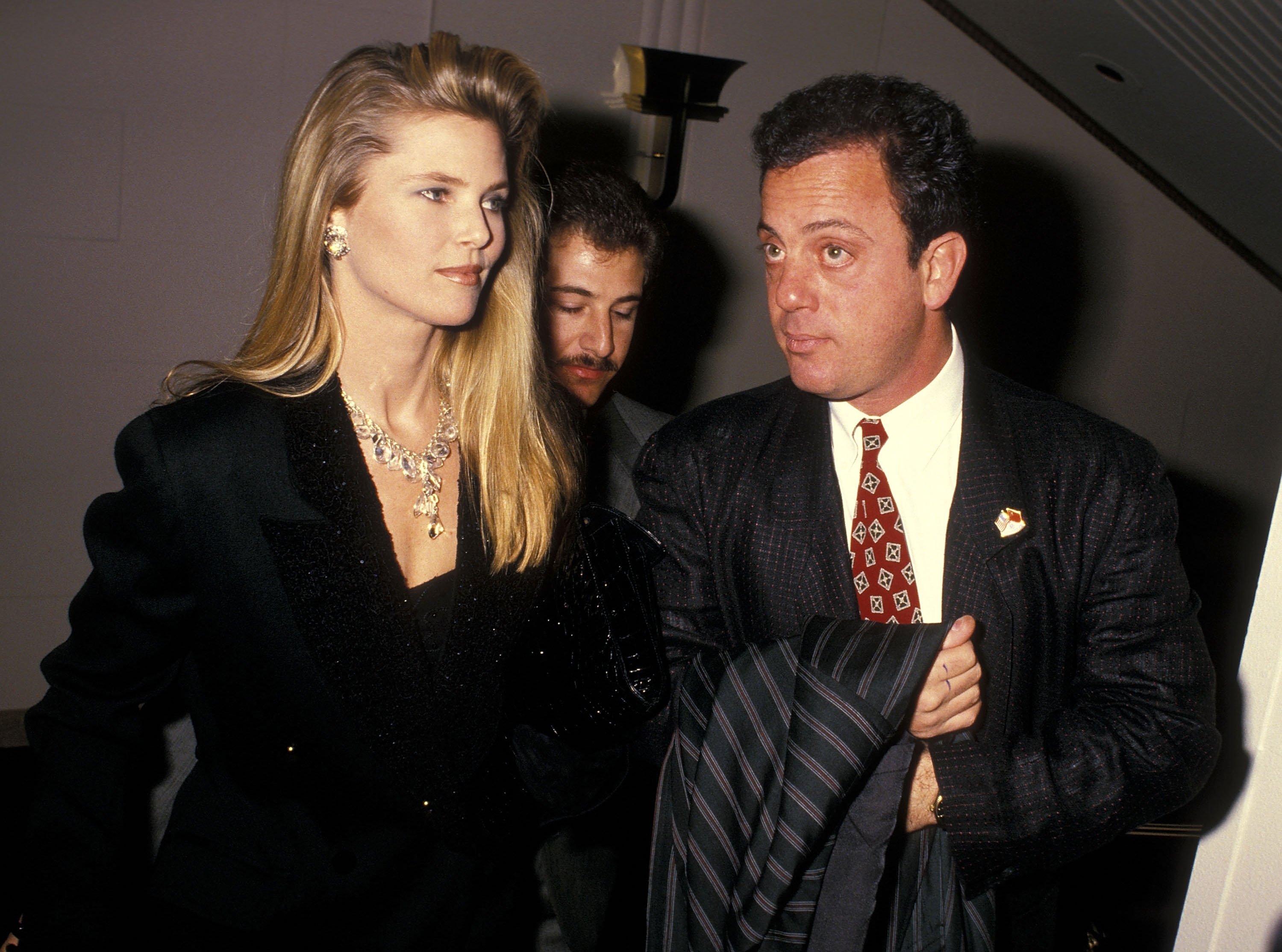 Two individuals in formal attire at an event; one wears a suit with a patterned tie, the other a tailored outfit with statement jewelry
