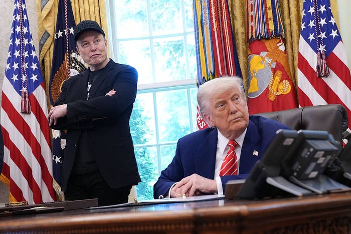 Two men in suits are in an office. One stands with arms crossed; the other sits at a desk, looking sideways. An official flag is visible behind them