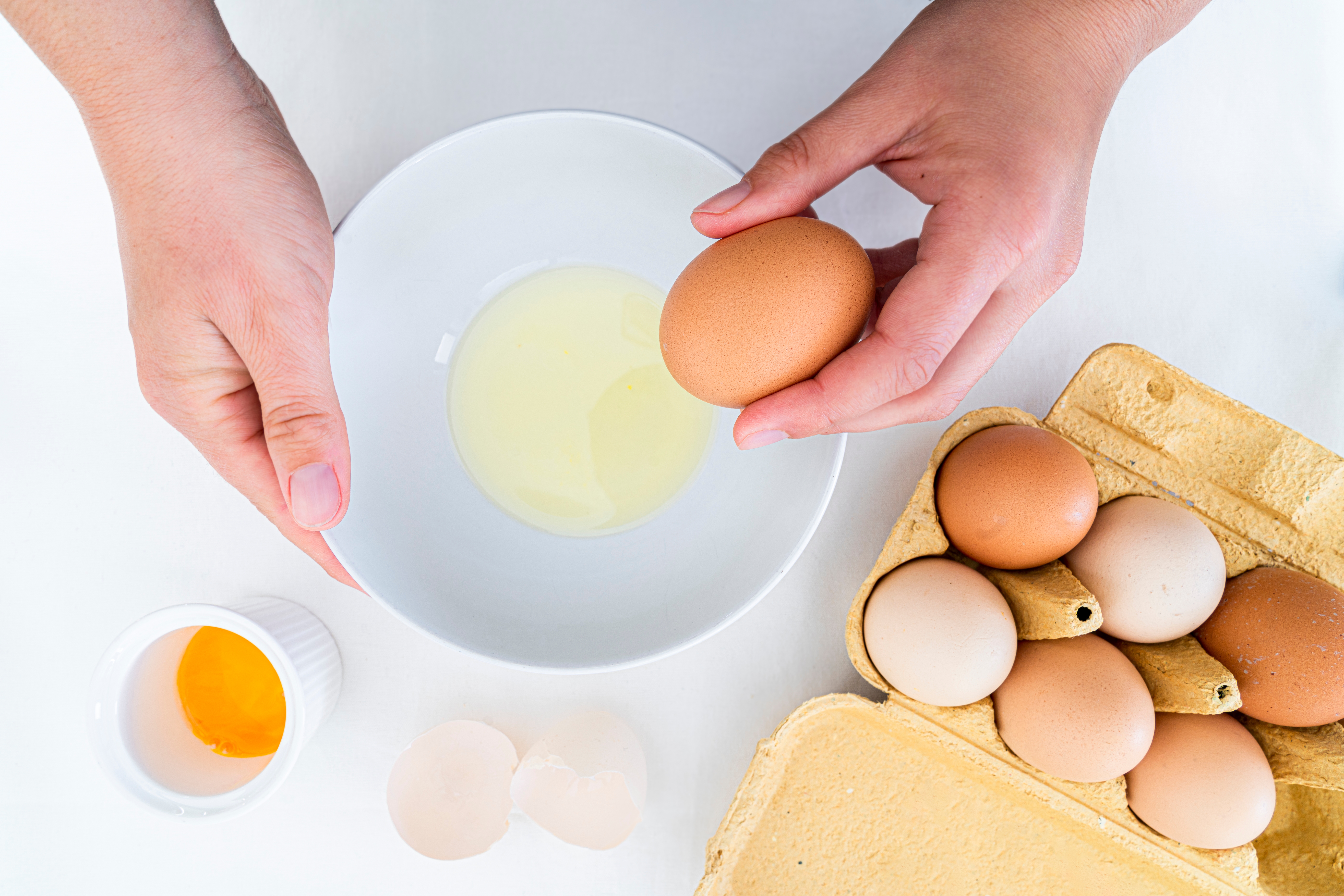 Hands cracking an egg into a bowl with an open egg carton and broken shell nearby