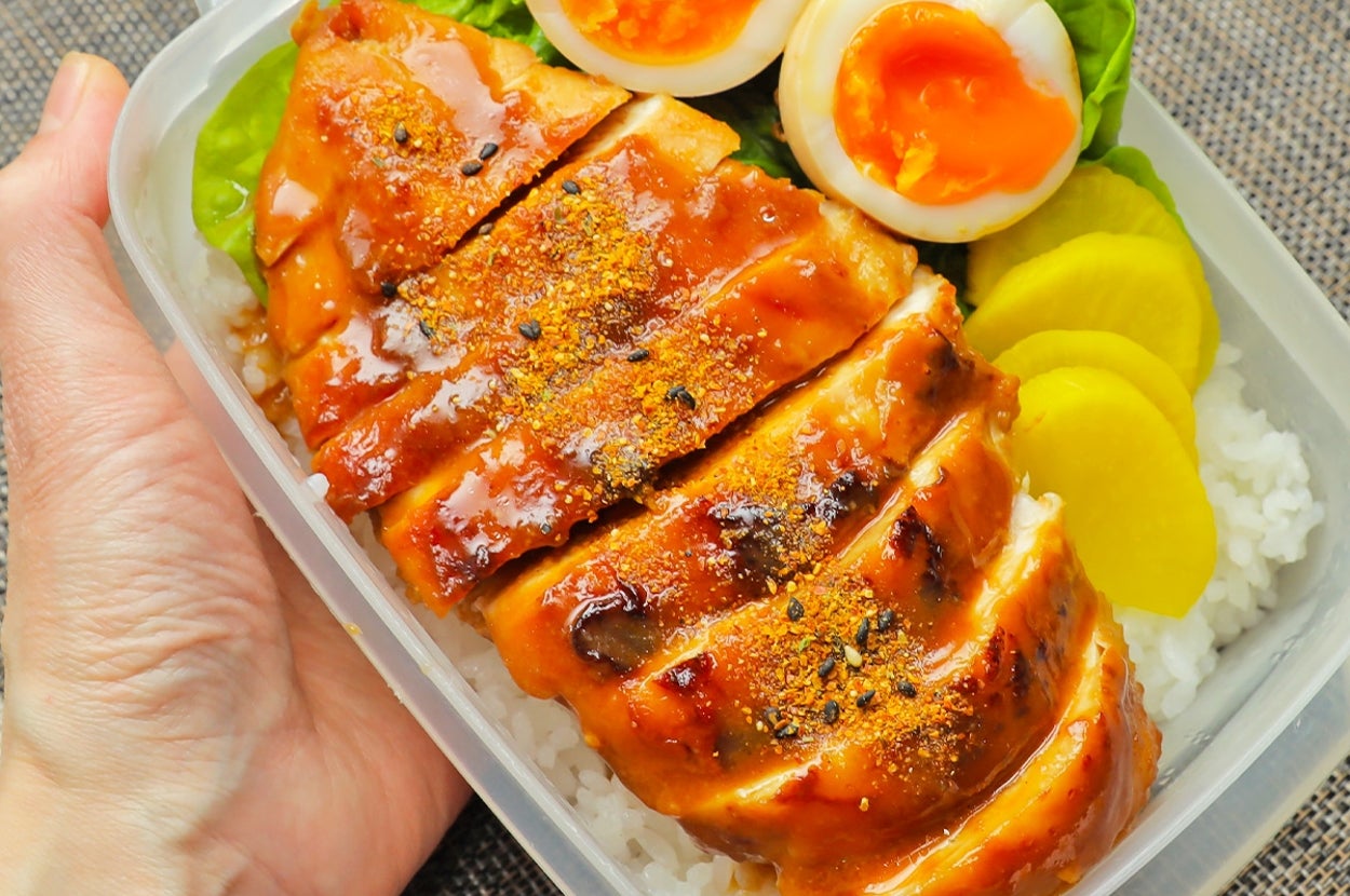 手作り弁当が手に持たれている。ご飯の上に照り焼きチキン、半熟卵、漬物、レタスがのっている。