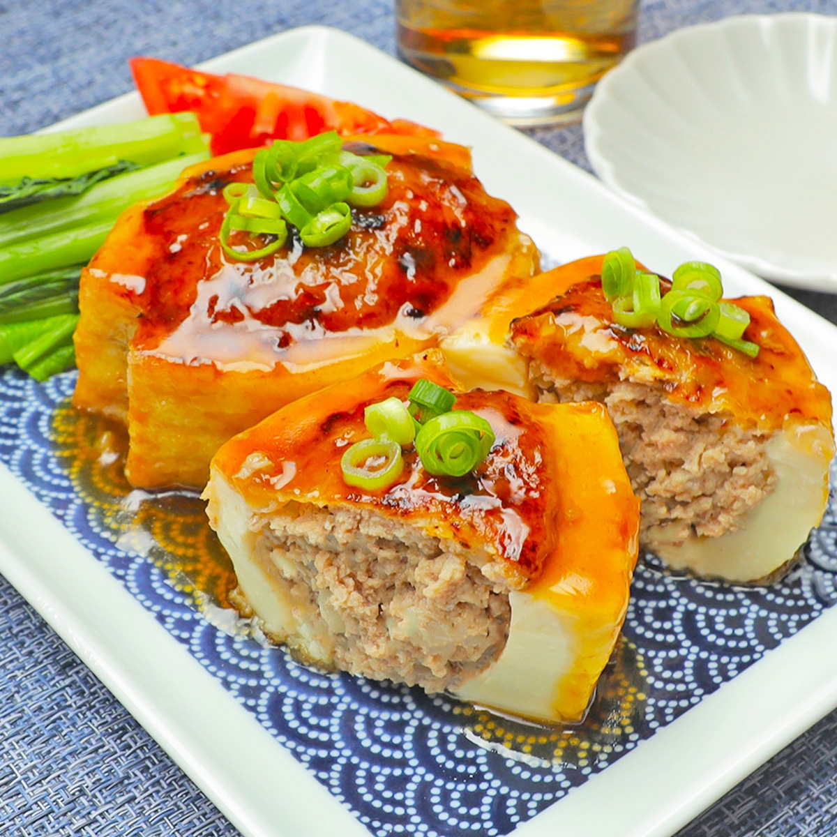 肉詰め厚揚げが皿に盛られ、ネギが散らされた料理。奥に飲み物と野菜が添えられている。