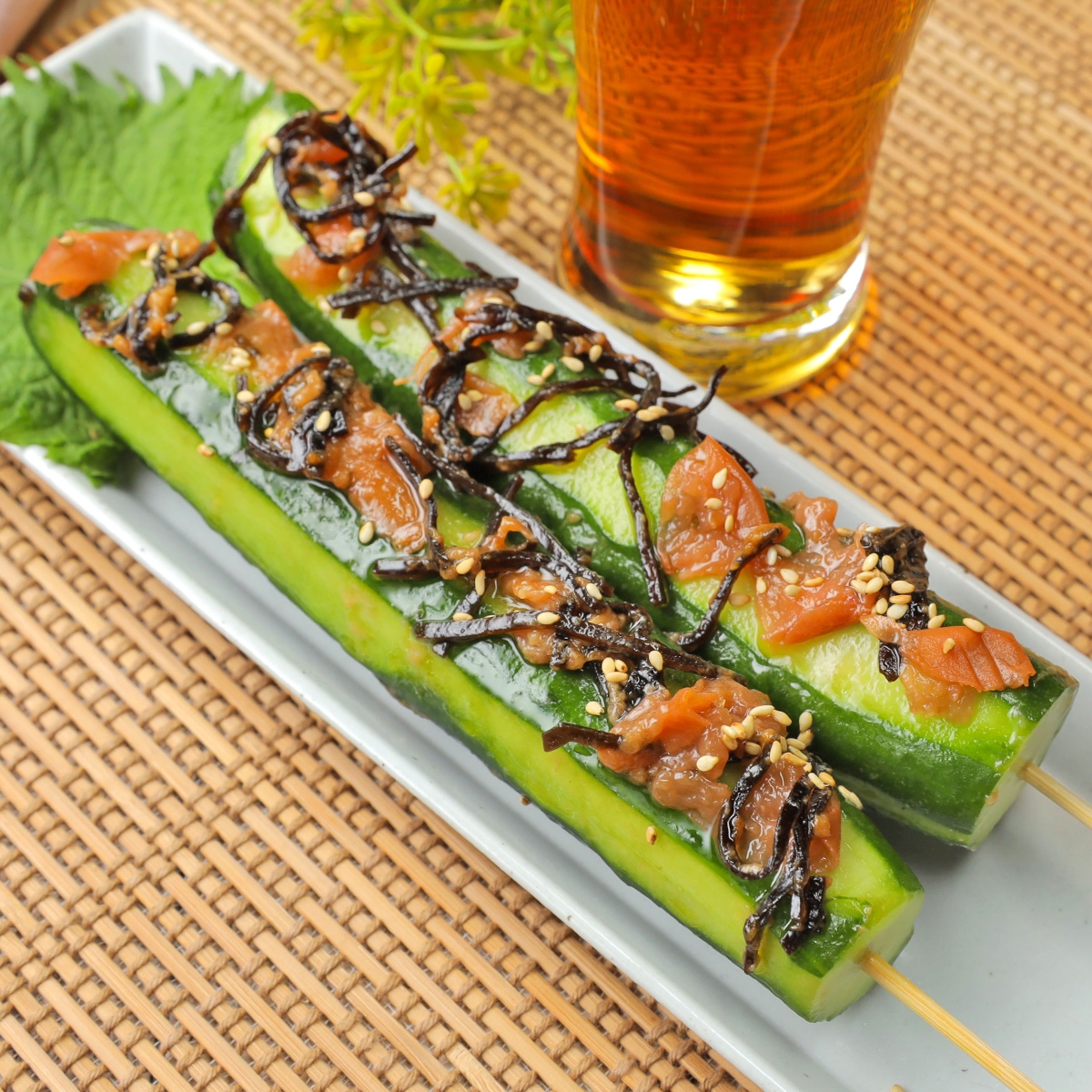 きゅうりに醤油漬けの昆布とゴマをトッピングした串料理が皿に乗っており、隣にはグラス入りの飲み物がある。