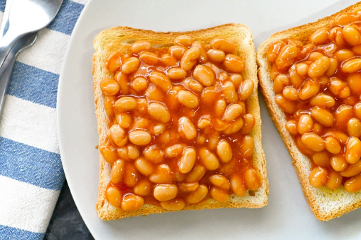 Two slices of toast topped with baked beans on a white plate, next to a striped cloth and cutlery