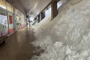 Outdoor shopping area with heavy snow piled against storefronts, partially blocking the walkway