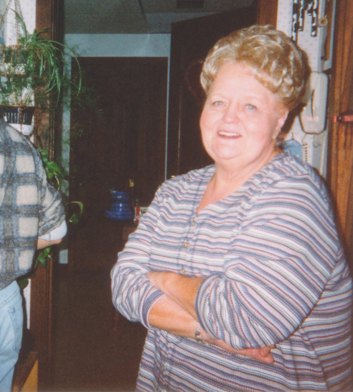 Person smiling indoors, wearing a striped top. Arms crossed in a relaxed manner, standing in a homely room setting