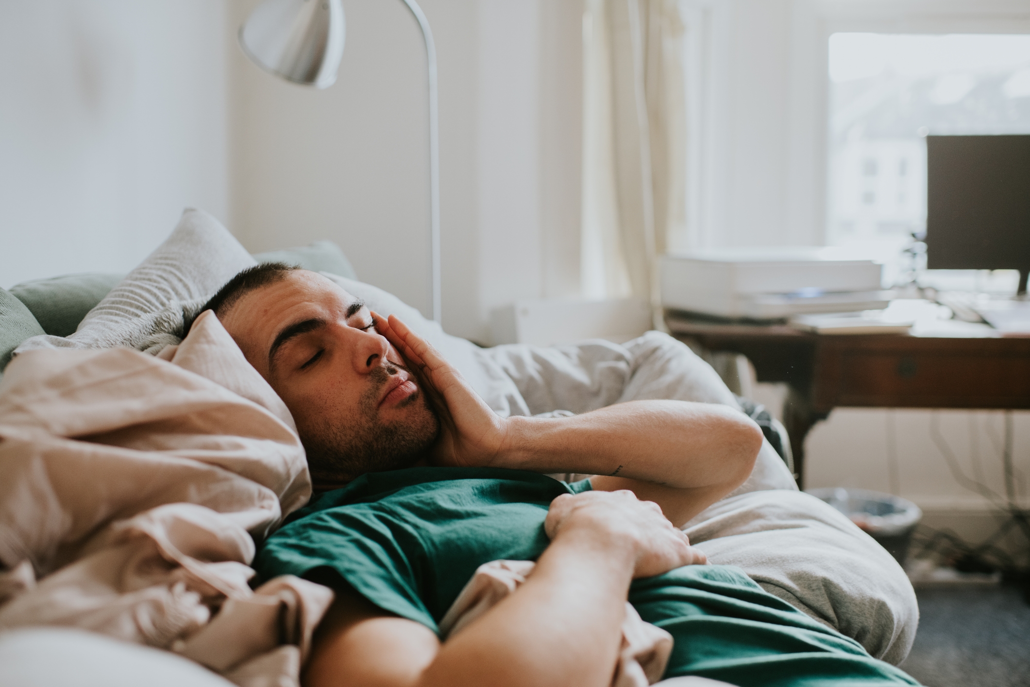 A person rests in bed with their hand on their face, suggesting contemplation or fatigue, in a home office setting with a desk and computer nearby