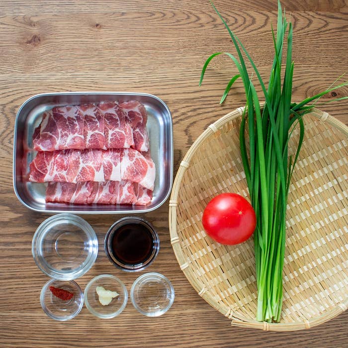 調理用の材料が木製テーブルに並べられている。金属トレーには薄切りの牛肉、籠にはトマトとネギの束があり、調味料が小皿に入っている。