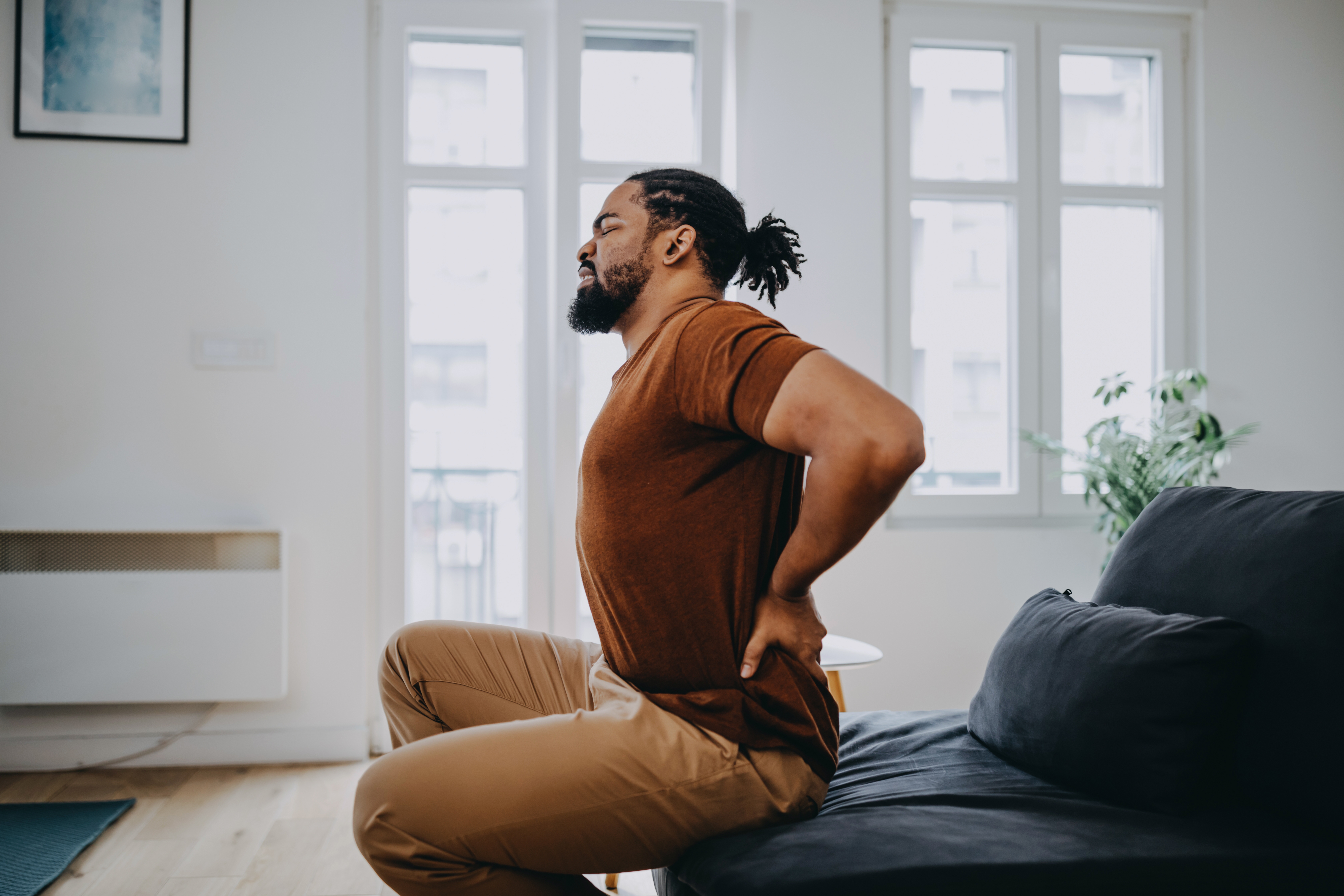 Personen som sitter på en sofa, strekker seg med hendene på korsryggen, og opplever sannsynligvis smertelindring. Naturlig lys fyller rommet fra store vinduer