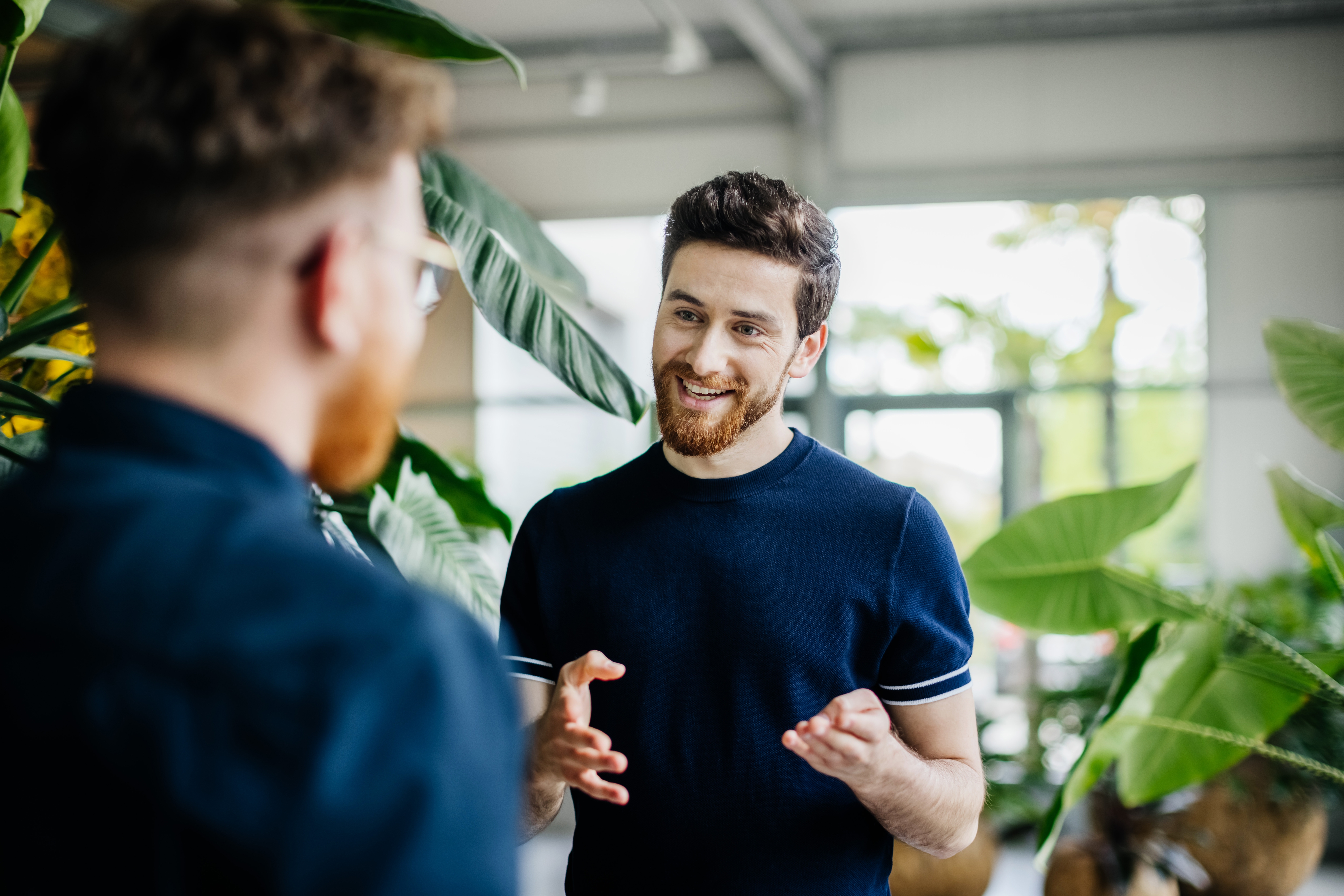 Mann smiler og gestikulerer energisk mens han snakker med en annen person i en frodig, plantefylt innendørs setting