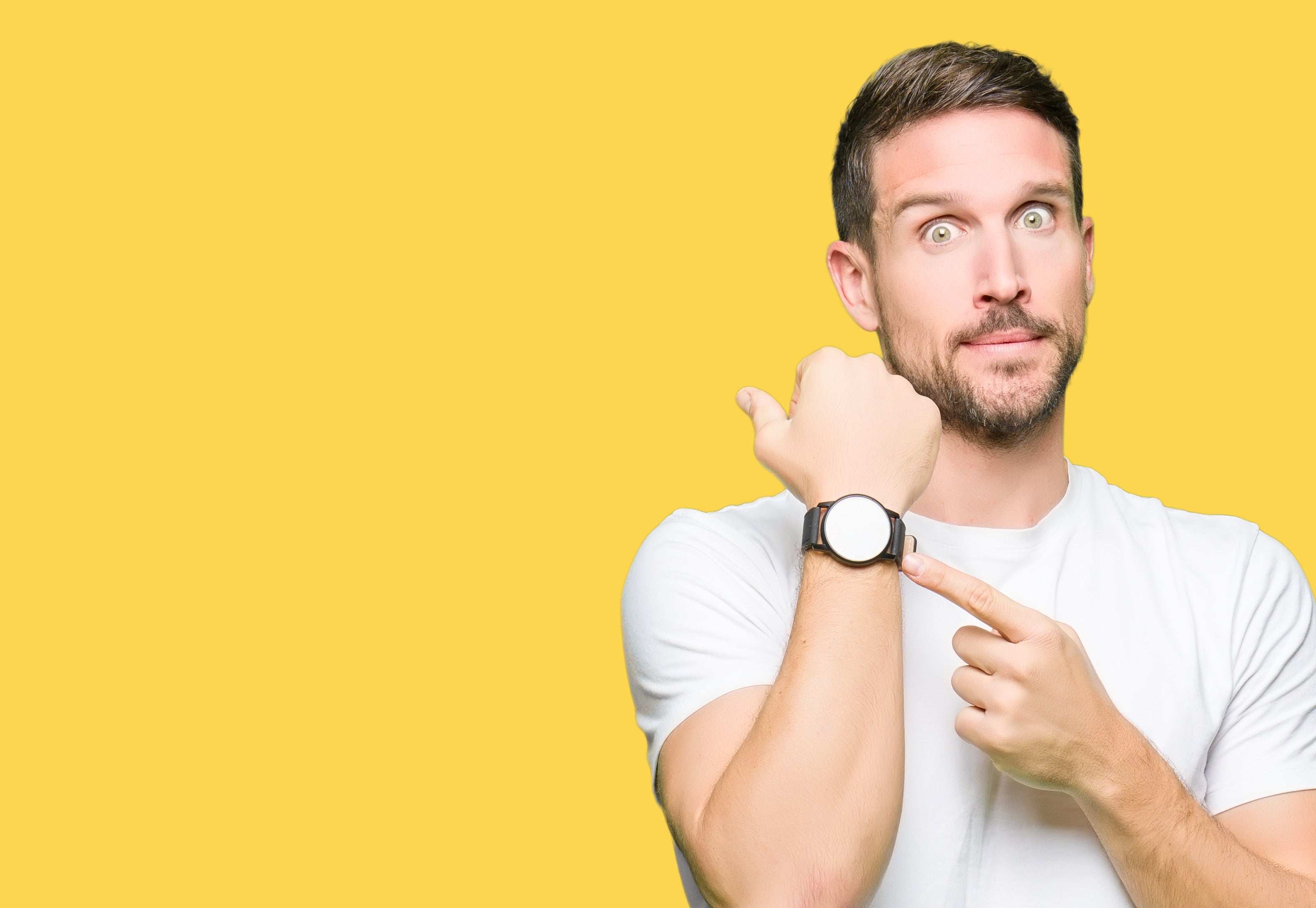 Mannen i en hvit t-skjorte ser overrasket ut mens han peker på en klokke på håndleddet mot en vanlig bakgrunn