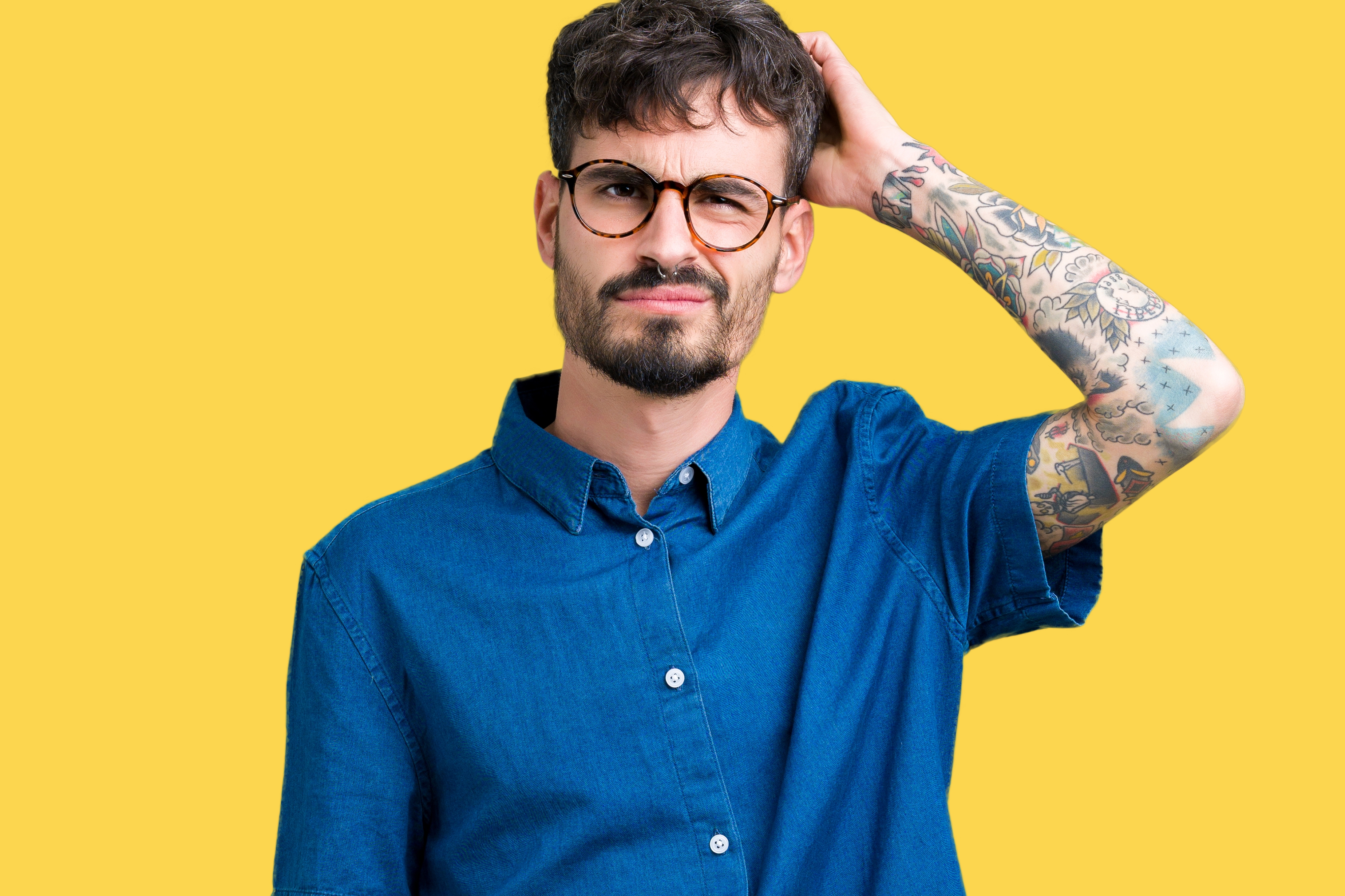 Mann med briller og tatoveringer på armen iført en denimskjorte med kort ermet, klør på hodet, på en vanlig bakgrunn