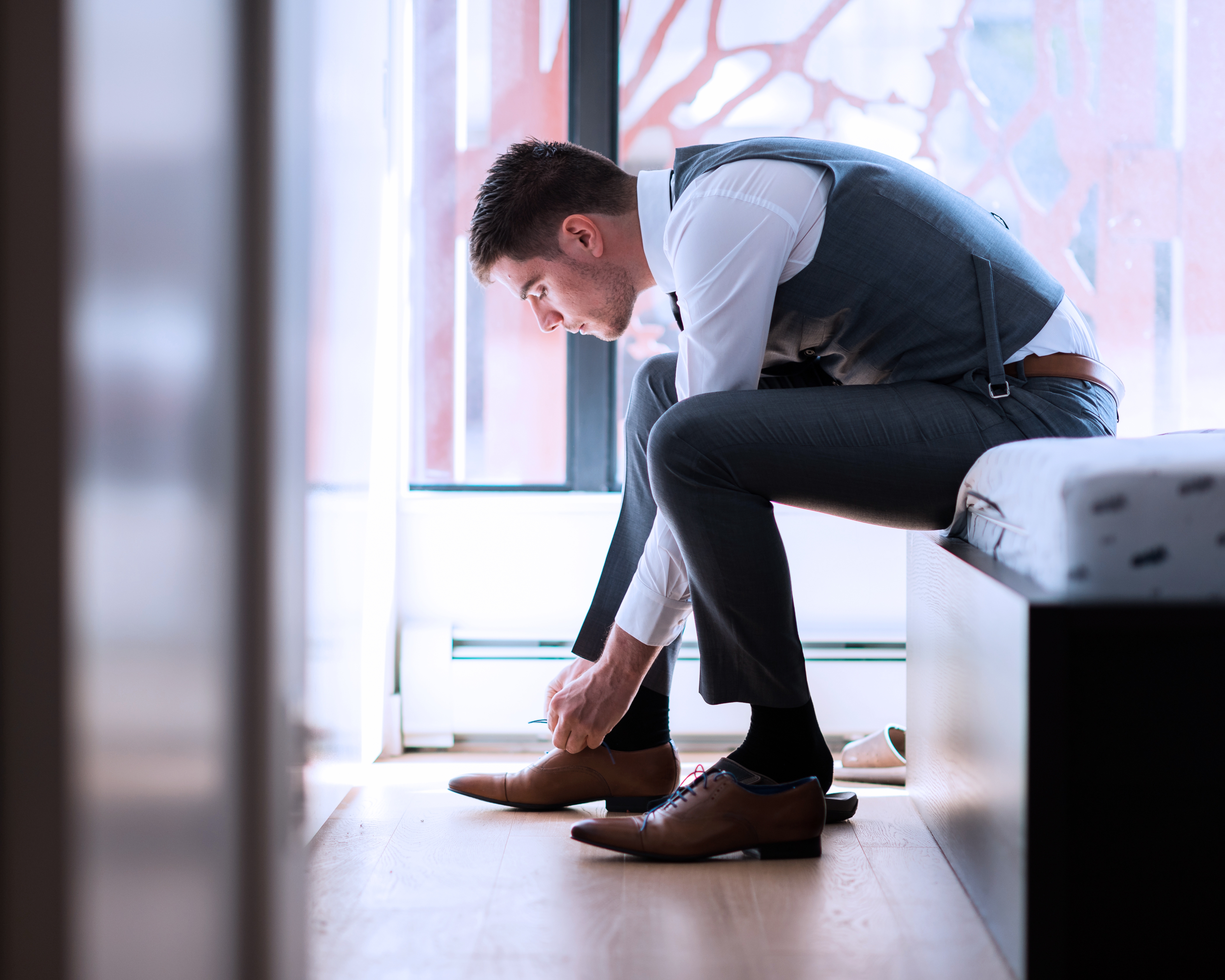 Mann i formell antrekk sitter på en seng og knytter klesskoene sine i et godt opplyst rom