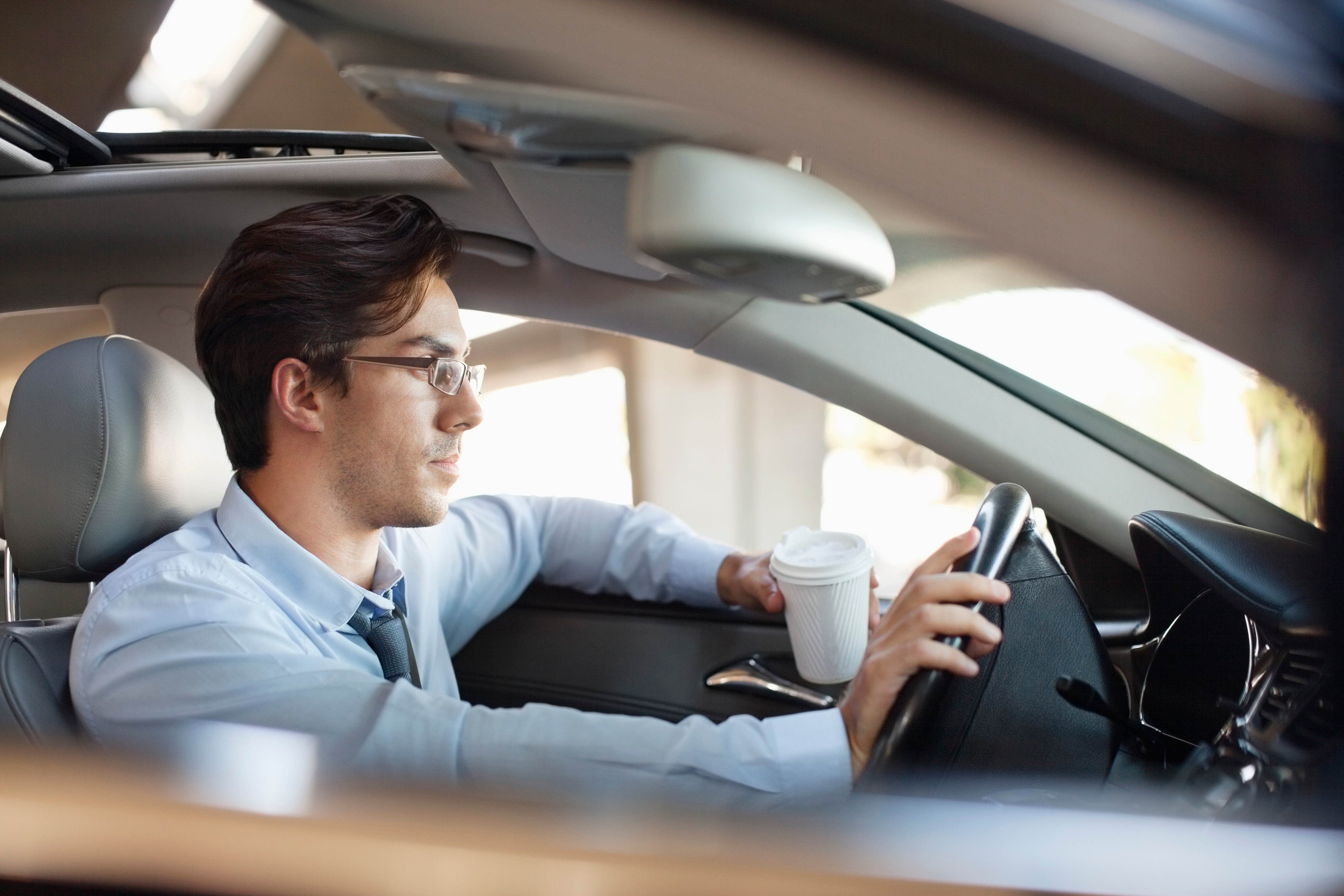 Mann i briller som kjører en bil mens du holder en kaffekopp, fokusert på veien foran. Han har på seg en formell skjorte og slips