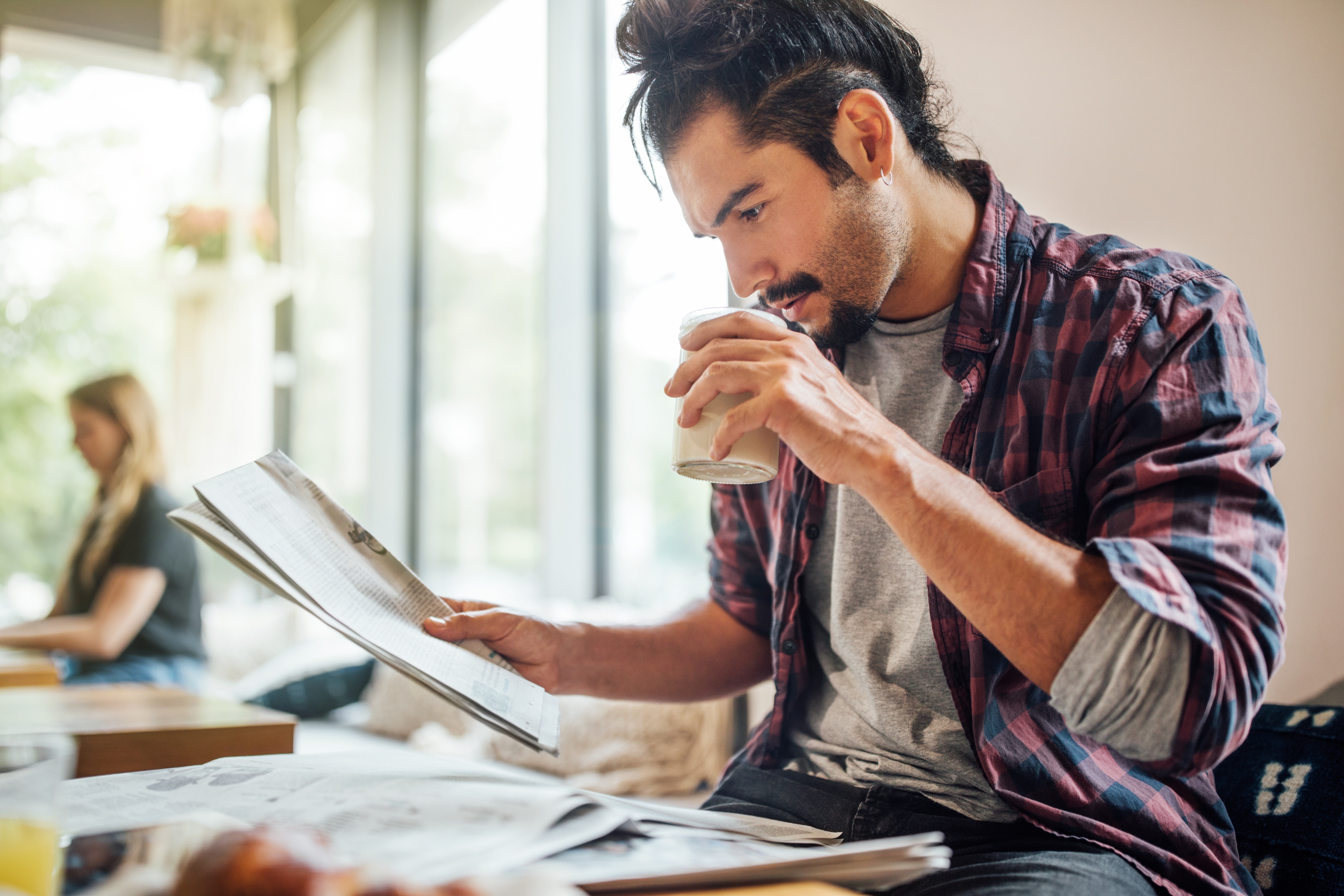 En mann med langt hår nipper til en drink og leser en avis på en koselig kafé, mens en kvinne sitter i bakgrunnen