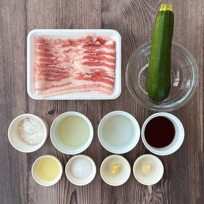 長いスライスの豚バラ肉、ズッキーニ、調味料が並んだ料理の準備風景。