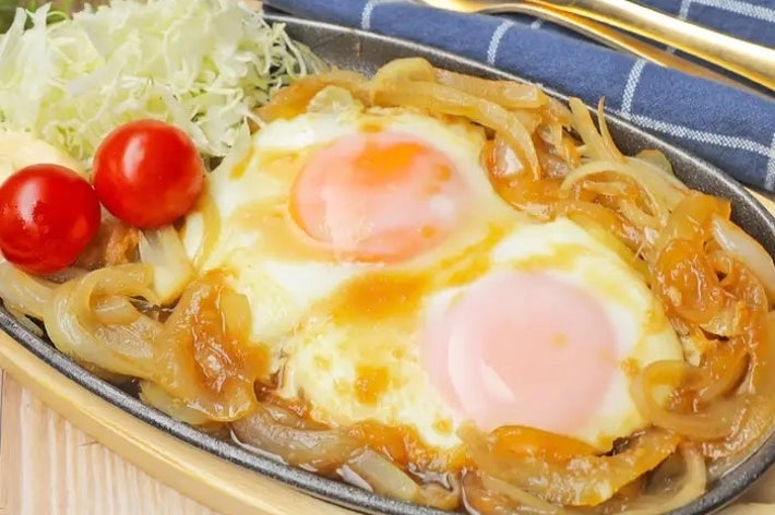 目玉焼きが玉ねぎと一緒に鉄板に乗せられ、キャベツとミニトマトが添えられた料理。チェック柄のナプキン。