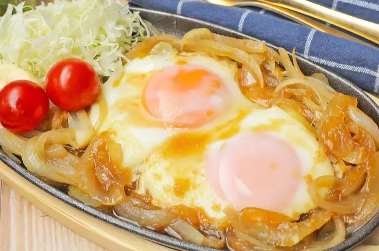 目玉焼きが玉ねぎと一緒に鉄板に乗せられ、キャベツとミニトマトが添えられた料理。チェック柄のナプキン。