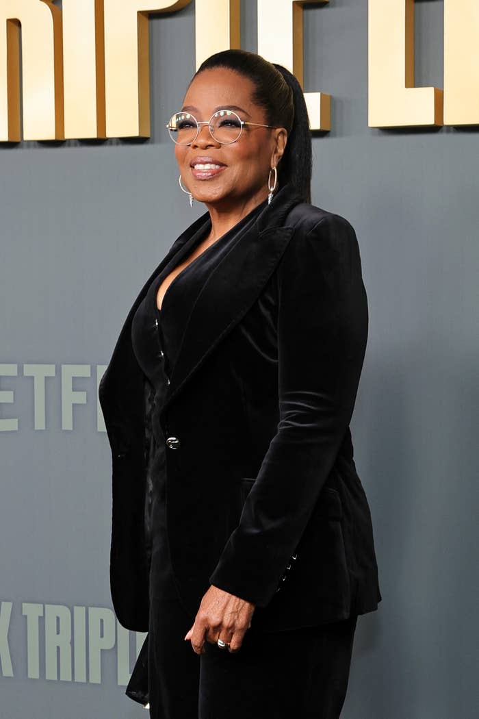 Oprah Winfrey smiling on a red carpet, with hoop earrings and round glasses