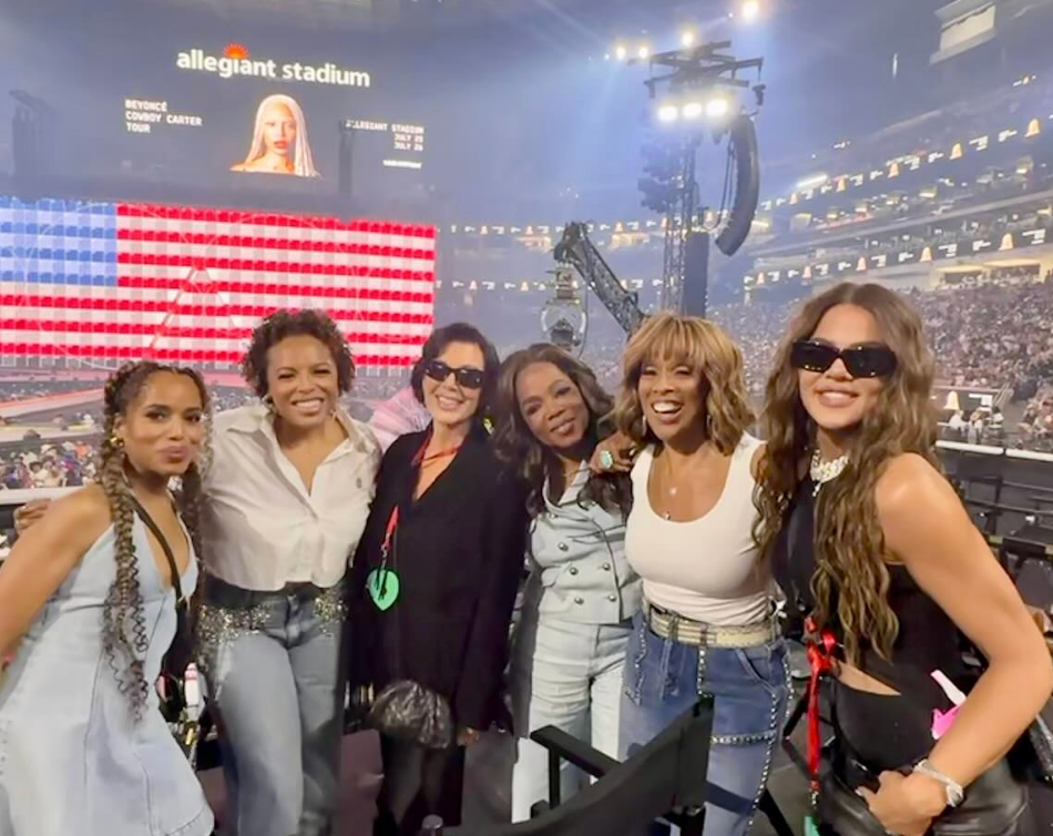 Six women pose for a photo at a concert with a large American flag display in the background. They're dressed in stylish, casual outfits