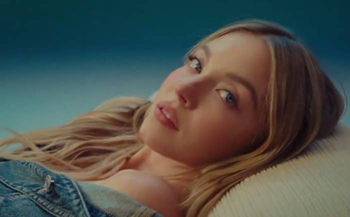 Person with long hair lies on a textured surface, wearing an off-the-shoulder denim top, gazing at the camera
