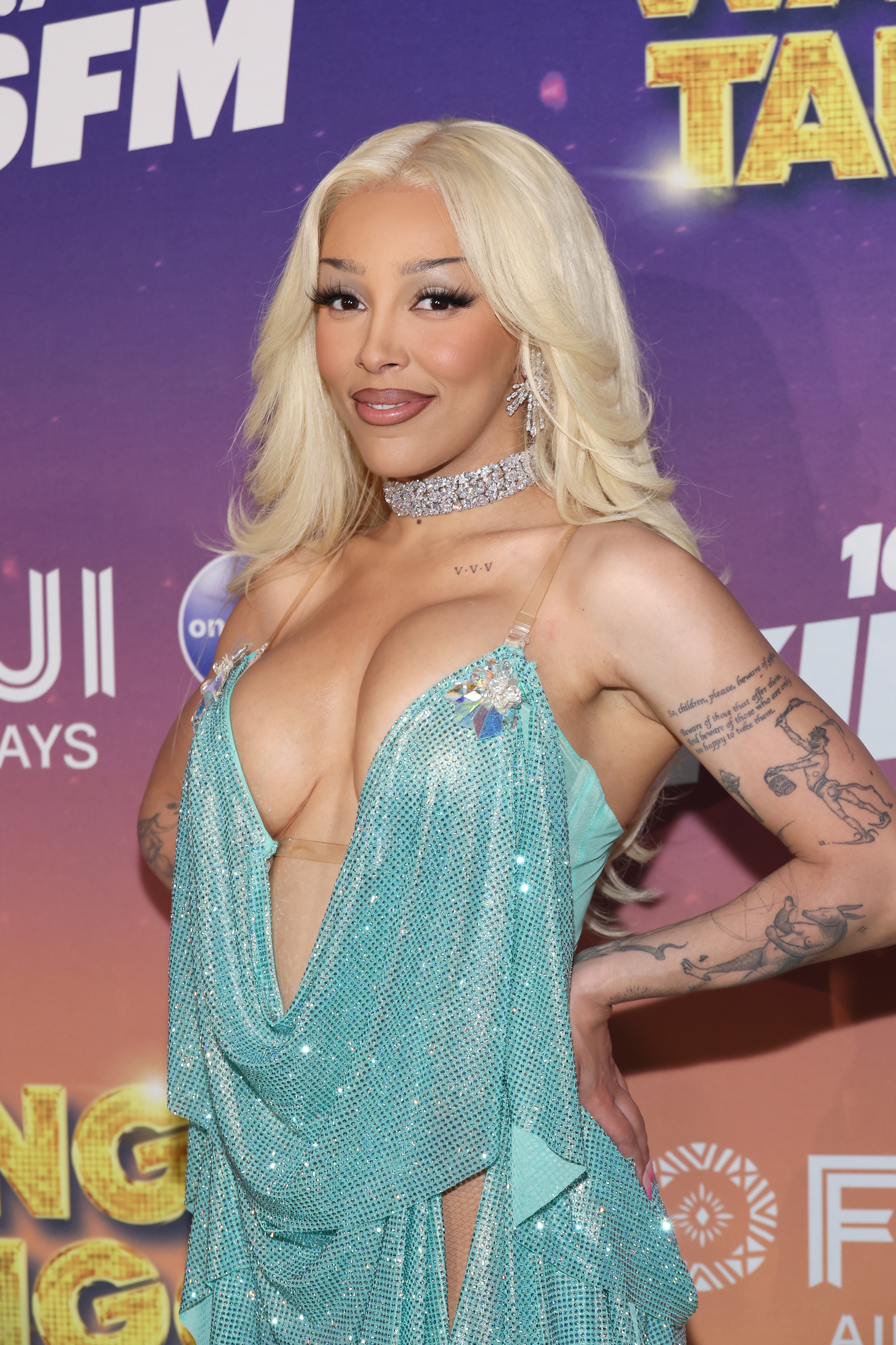 A person poses on the red carpet in a sparkling, deep V-neck dress with a choker necklace, displaying tattoos on their arms