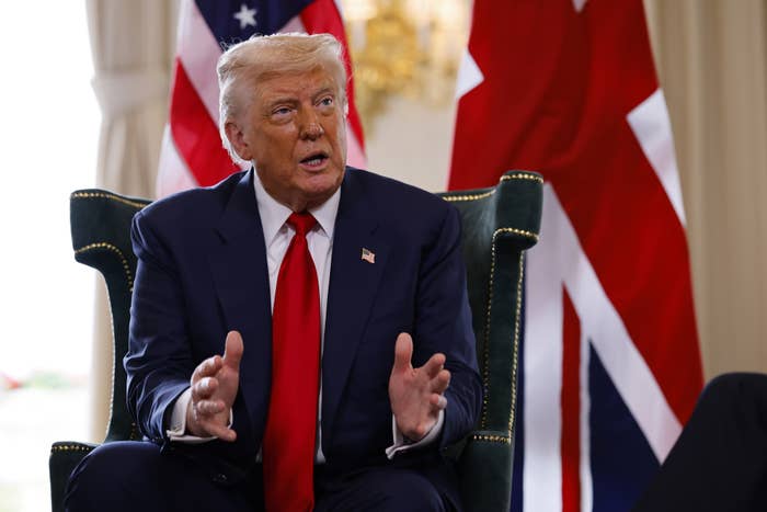 A person in a suit with a red tie is speaking, seated in front of flags