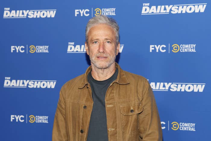 A person at a promotional event for &quot;The Daily Show,&quot; wearing a casual jacket over a t-shirt, standing in front of a branded background