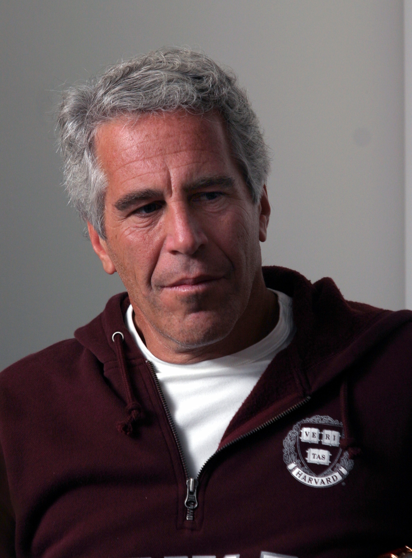A person with short gray hair wearing a dark hoodie and white shirt looks thoughtful indoors