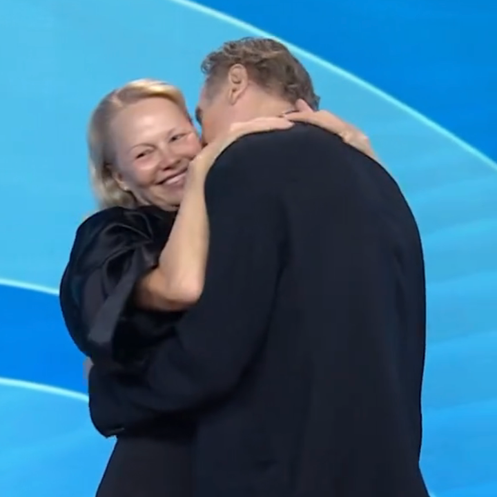 Two people embrace joyfully on stage during a televised event. The person on the left smiles brightly at the audience. Text reads: “THEY’RE HERE!”