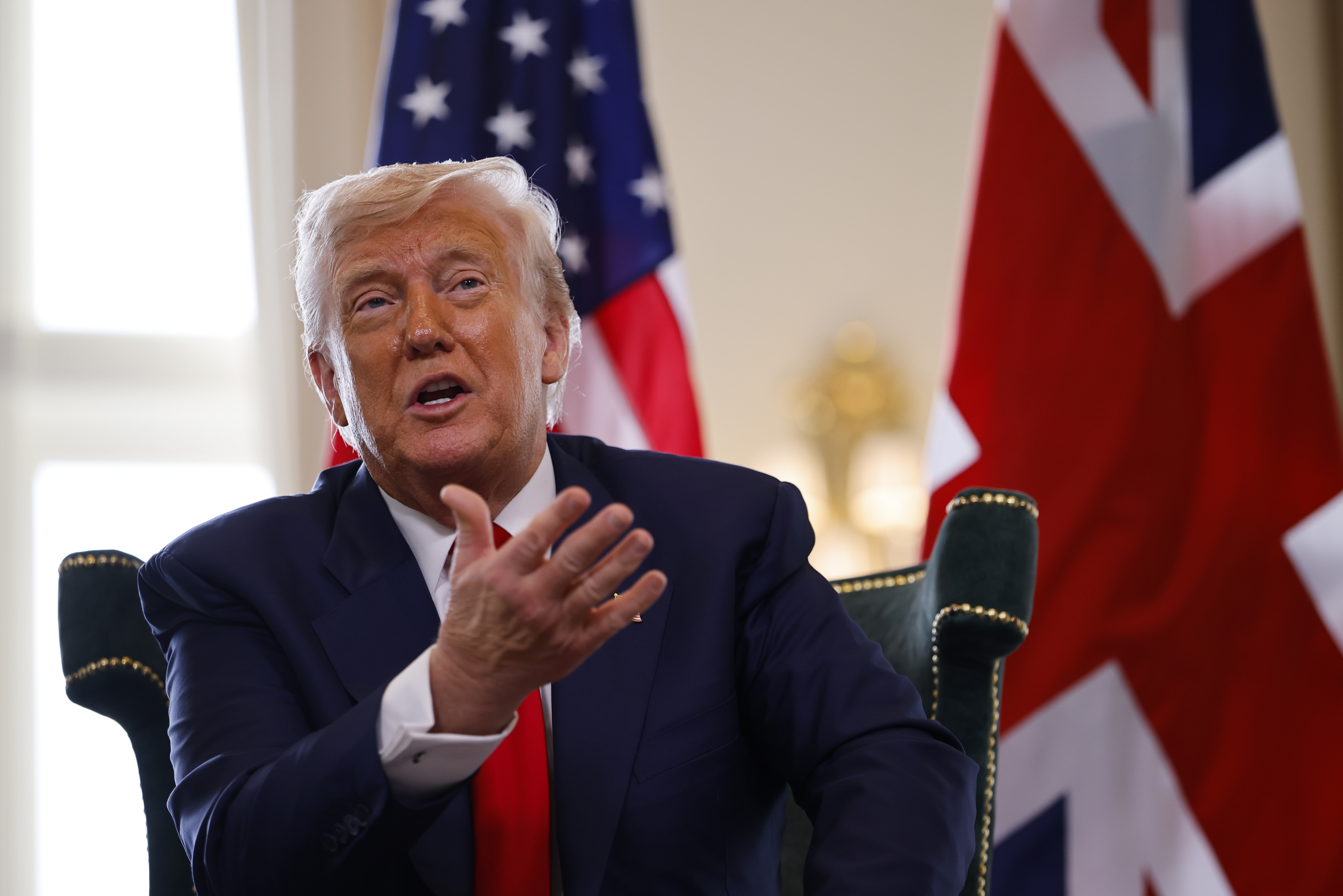 A person in a suit gestures while sitting in front of American and Union Jack flags