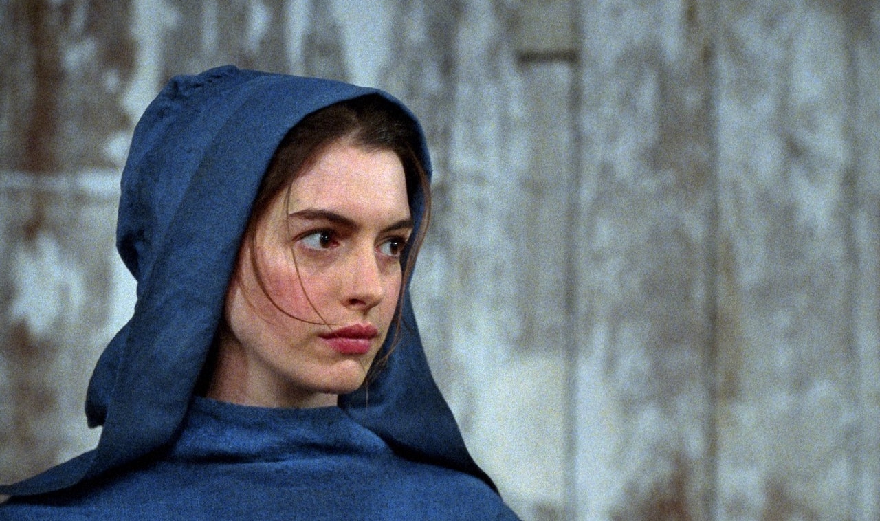 Person in simple hooded costume looking to the side against a weathered backdrop, seemingly from a historical or dramatic film scene