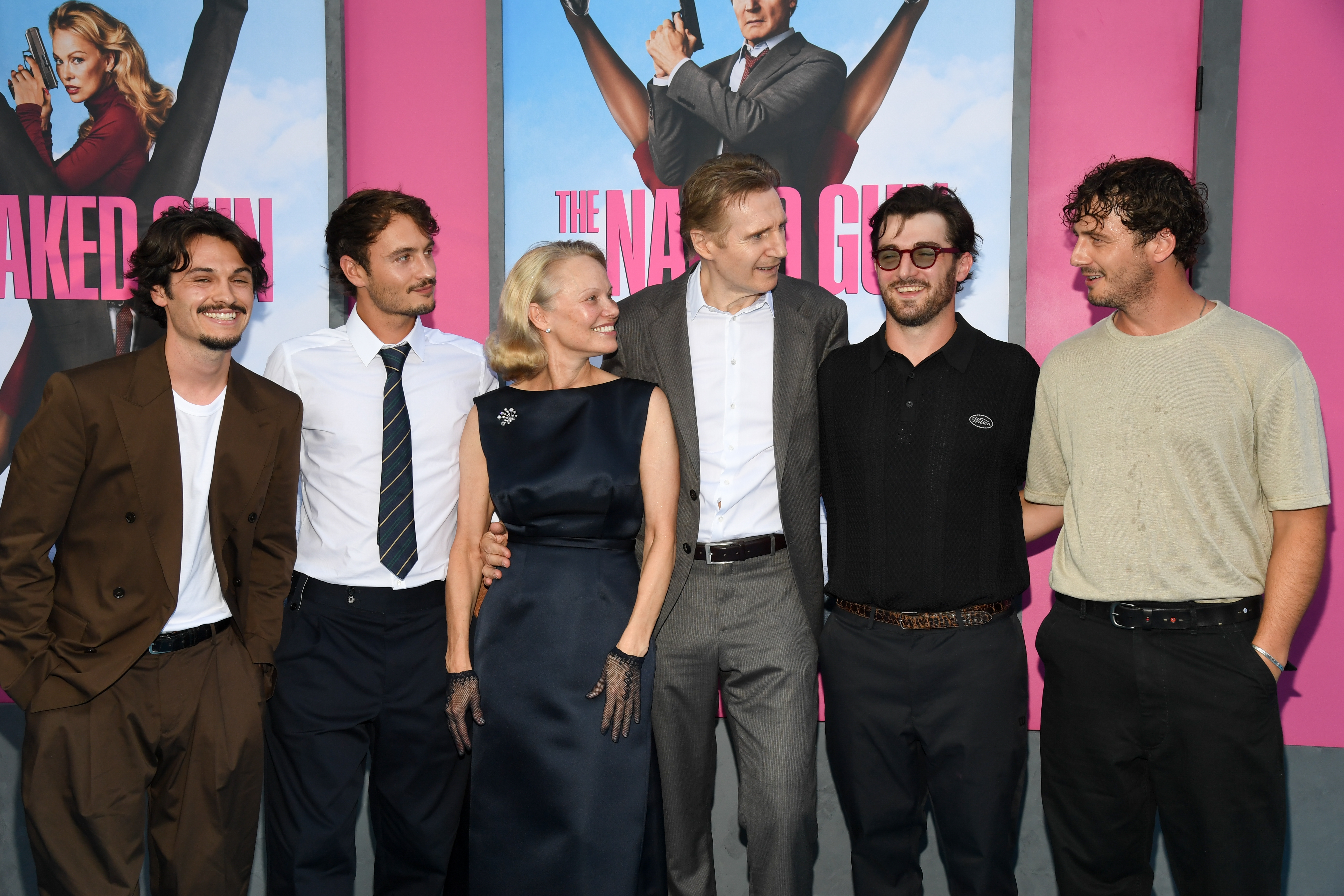 A group of six people smiling and chatting at a movie premiere, standing in front of "The Naked Gun" posters