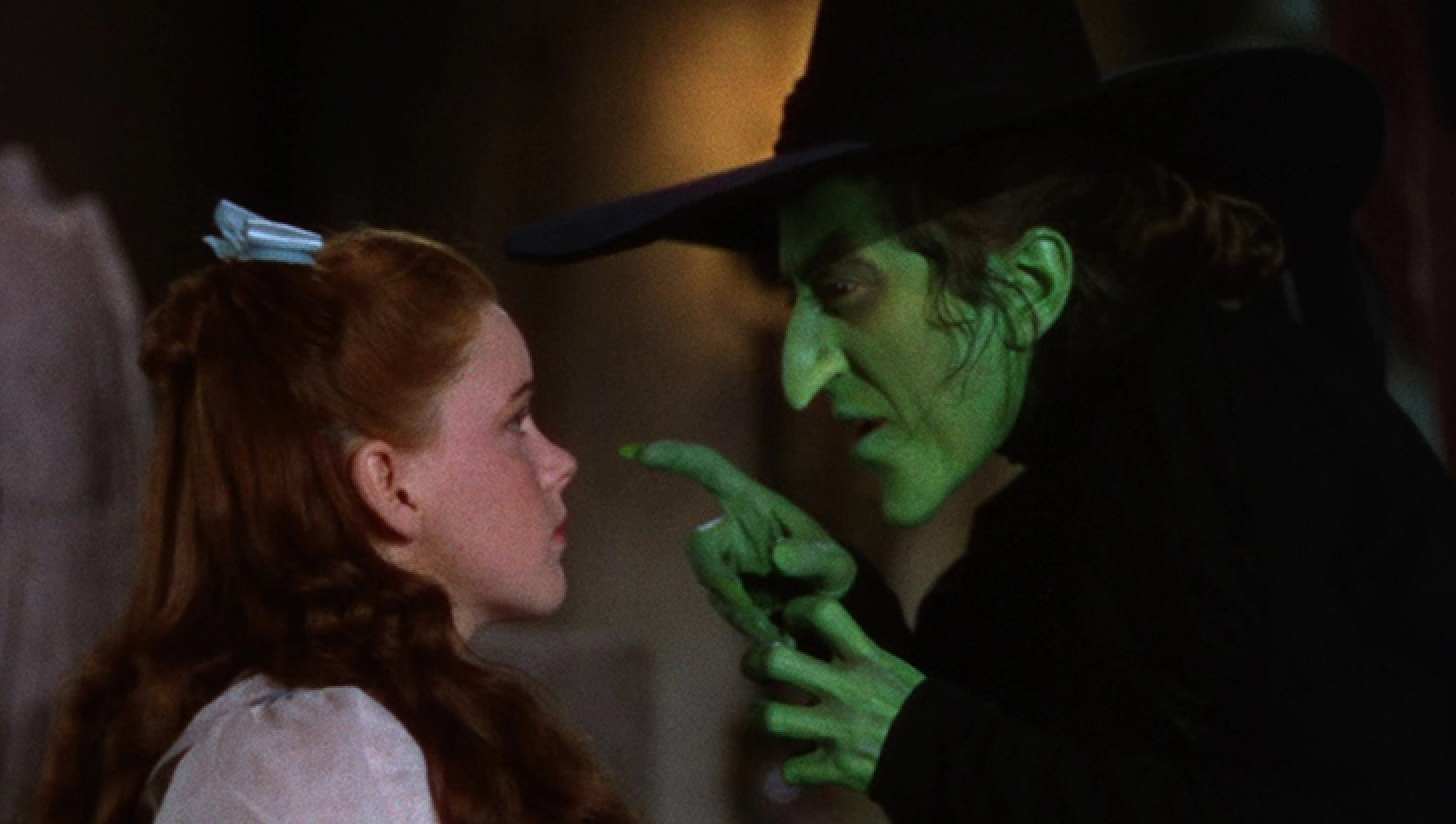 A woman in a witch costume with a pointed hat and green makeup menacingly points at a young girl with braided hair, in a scene from a film