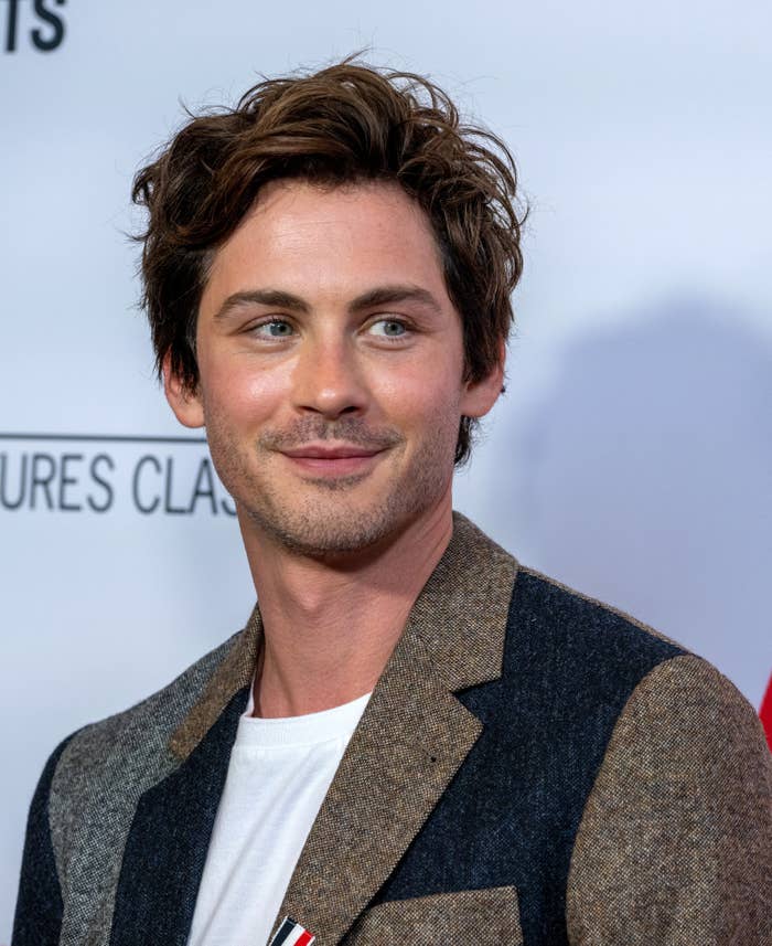 Logan Lerman with short, tousled hair in a textured blazer and white shirt on a red carpet smiling slightly to the side
