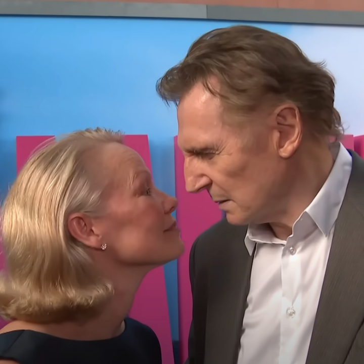 Two individuals stand close together at a red carpet event. The man wears a suit, and the woman is in an elegant dress