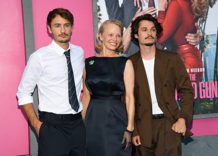 Three people pose at an event, with one wearing a white shirt and tie, another in a dark gown, and the third in a brown suit