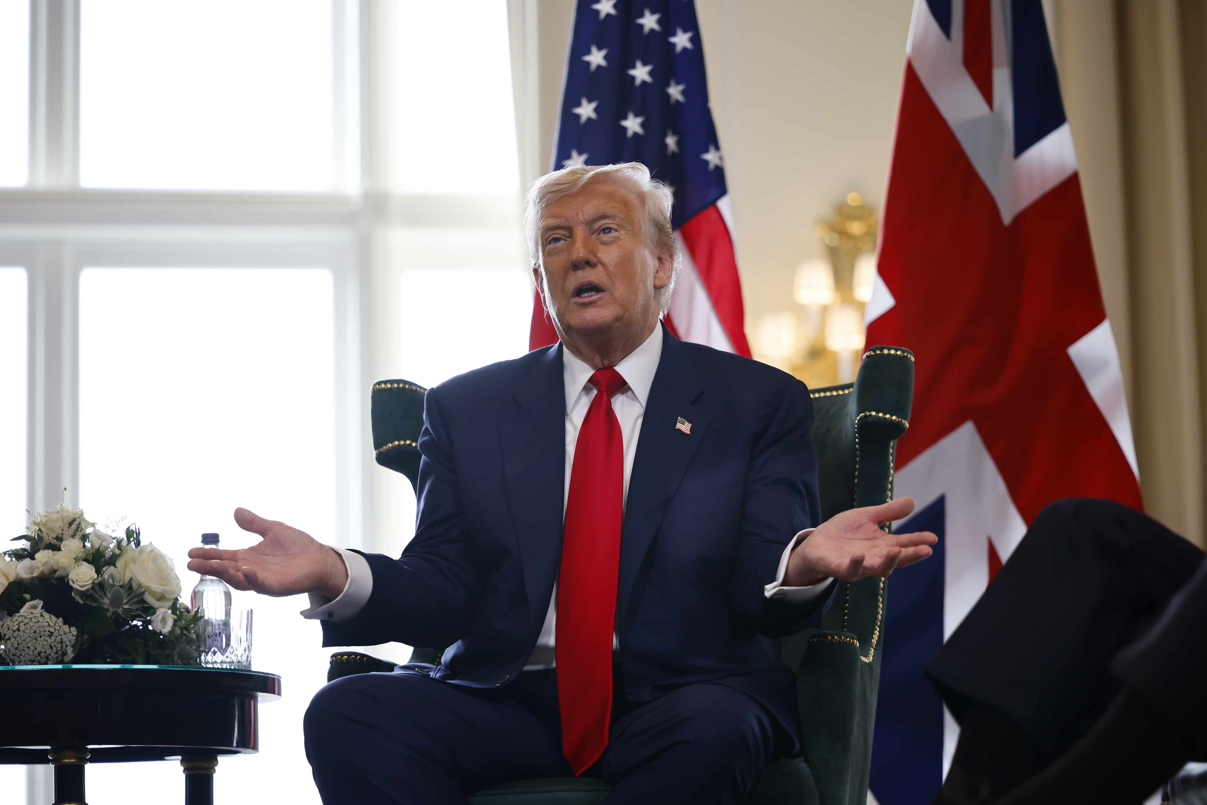 I don’t know who this person is, but they are seated, wearing a suit with a red tie, with U.S. and U.K. flags in the background