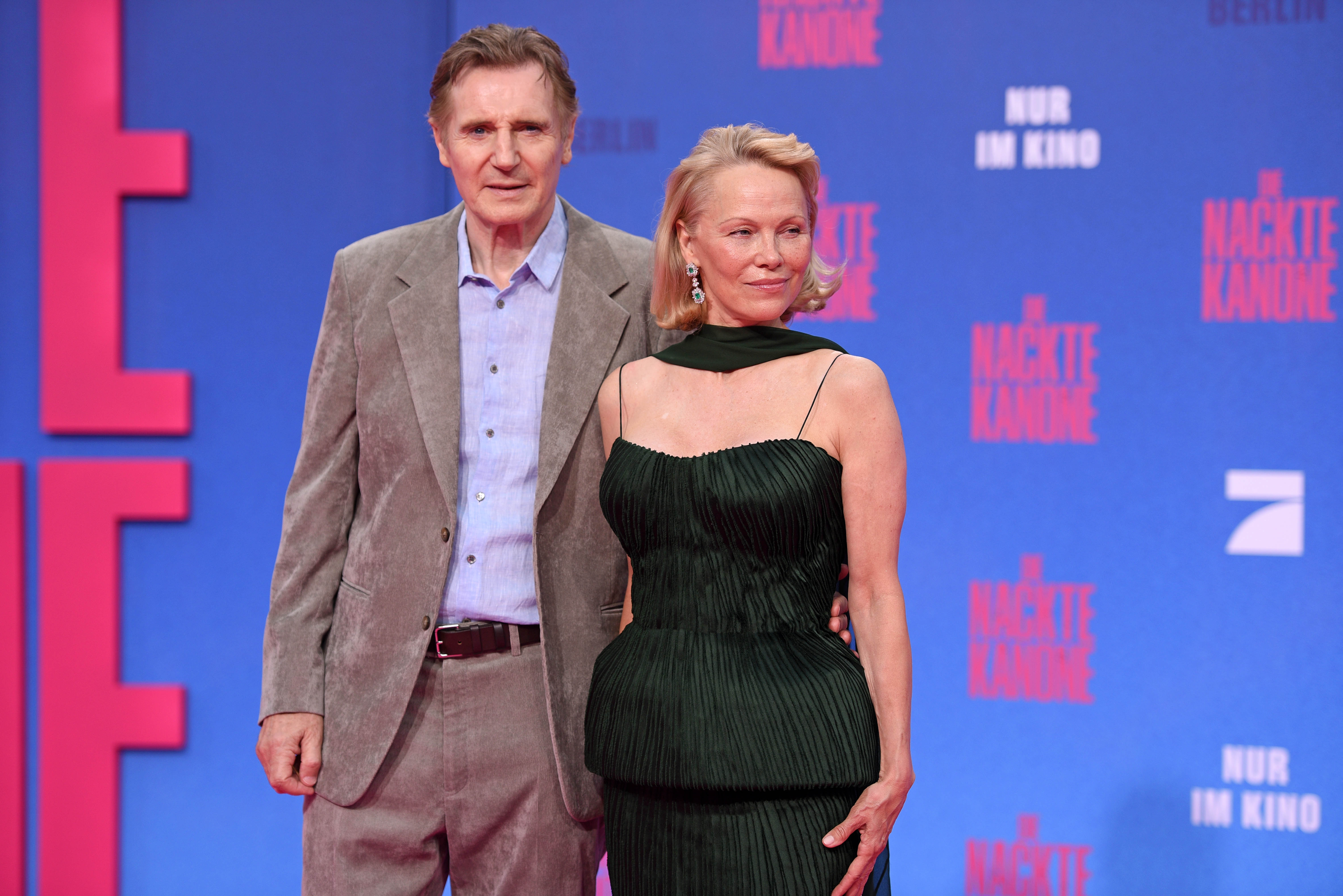 A man in a suit with no tie and a woman in a sleeveless, fitted gown on a red carpet