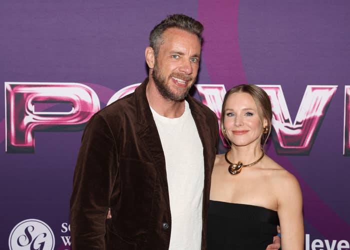 Dax Shepard and Kristen Bell smile on a red carpet. The man wears a velvet jacket over a white shirt; the woman wears a strapless dress and bold necklace