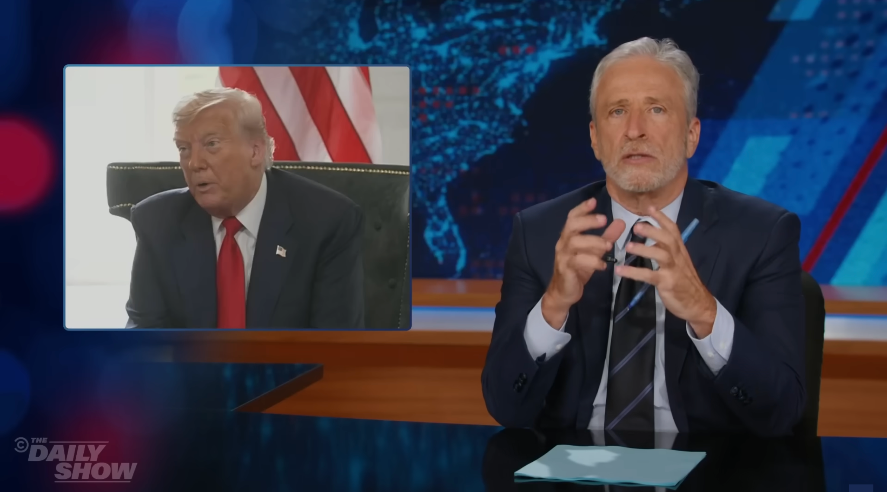 A person in a suit gestures while seated at a news desk, with an inset of another person in a suit appearing on a screen beside them