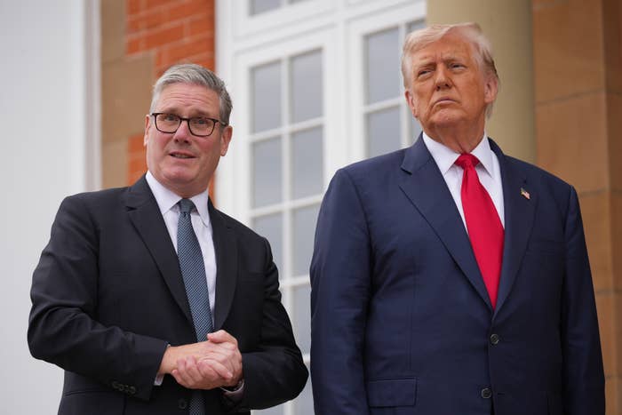 Two men in suits stand together outside a building with large windows; one wears a blue tie, the other a red tie